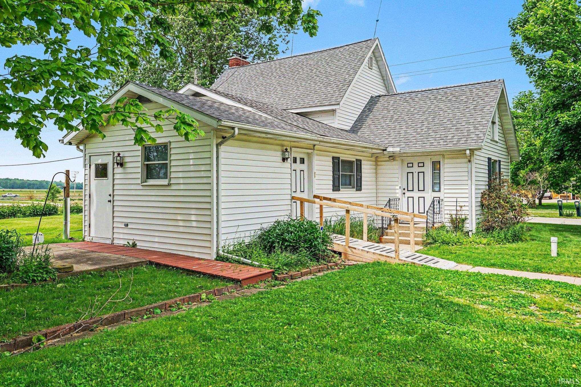White siding exterior featuring a gable roof, an accessible ramp leading to the main entrance, multiple windows with dark shutters, and a concrete patio