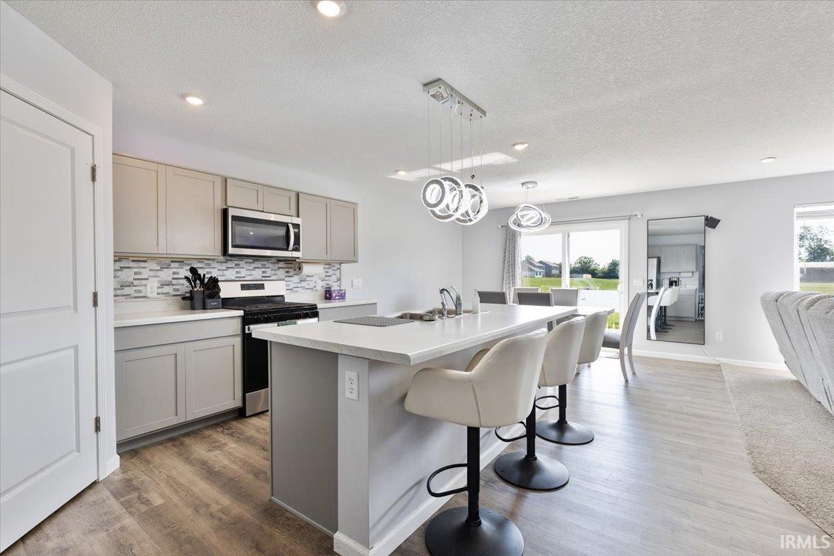 Open-concept kitchen featuring an island with a sink, light-toned cabinetry, stainless steel microwave, a tile backsplash, and wood-finish flooring