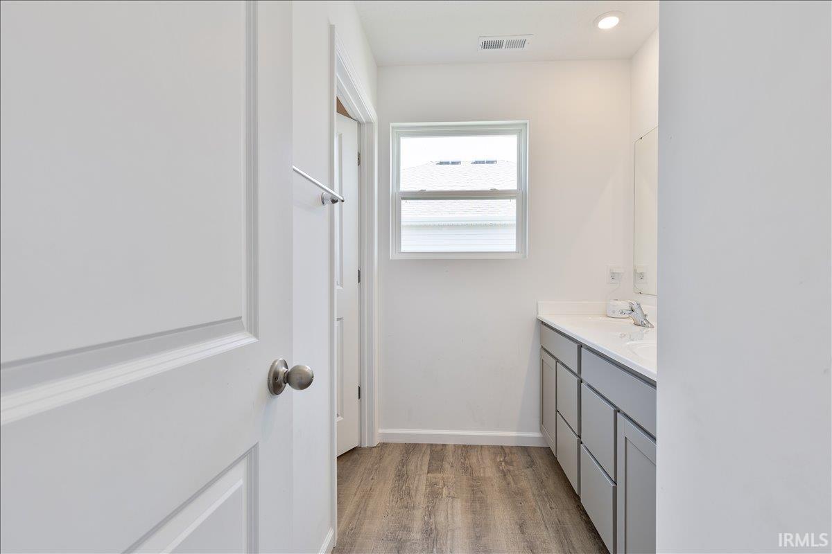 Bathroom with wood-finish flooring, a double vanity featuring white countertops, and a rectangular window with blinds