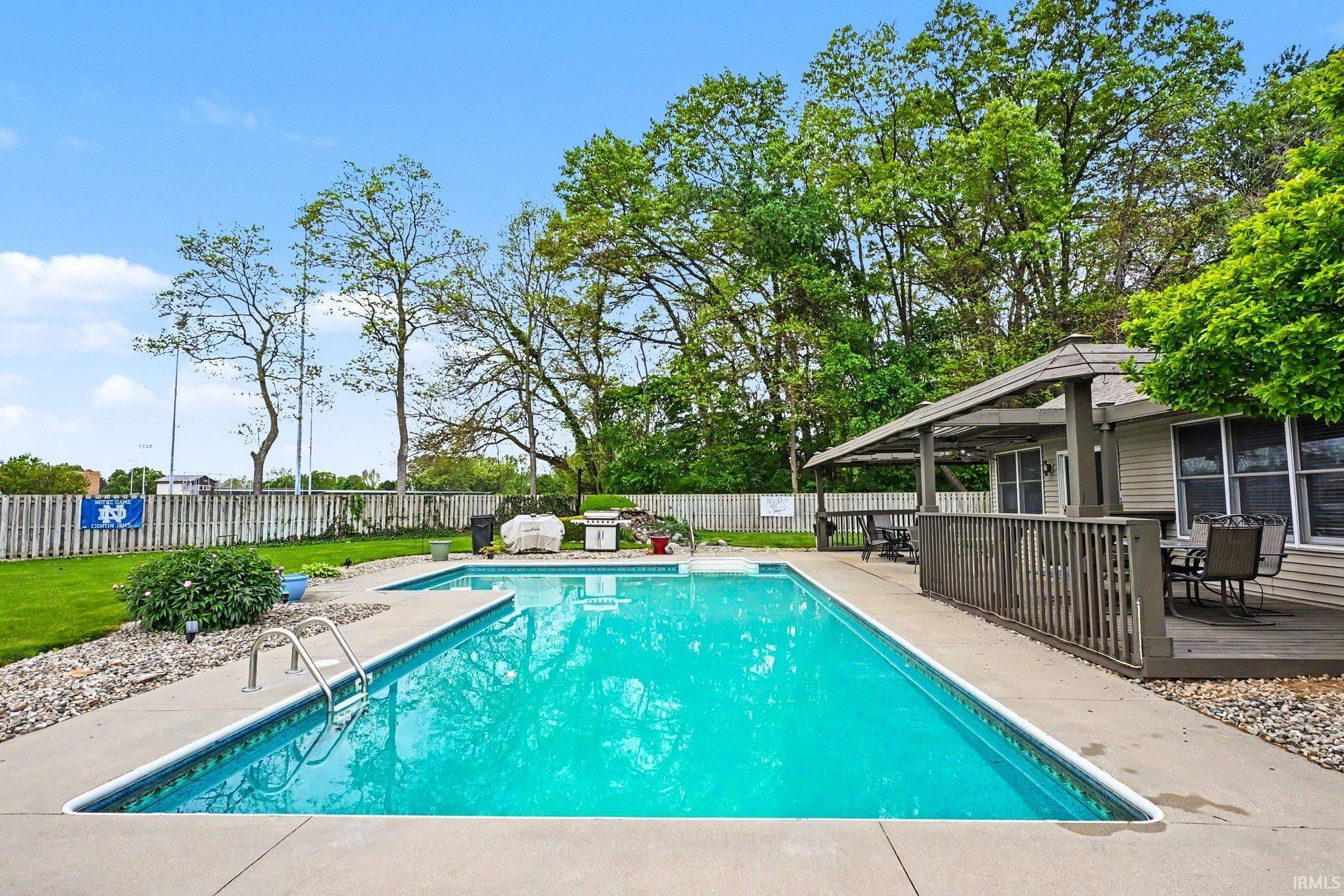 Inground swimming pool with a concrete surround, wood-tone deck, attached covered pergola, and a private perimeter fence