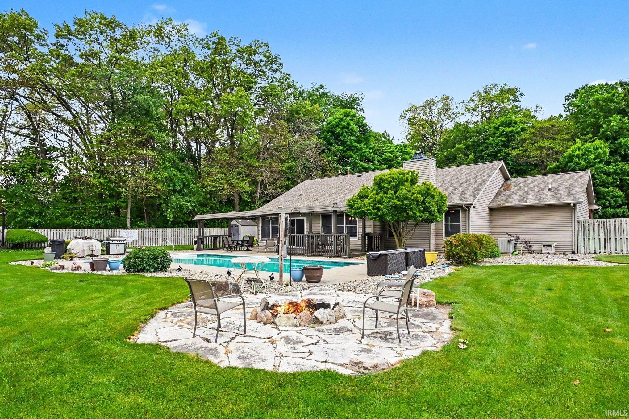 Expansive backyard featuring a stone fire pit area, a fenced swimming pool with a covered patio, and mature perimeter trees
