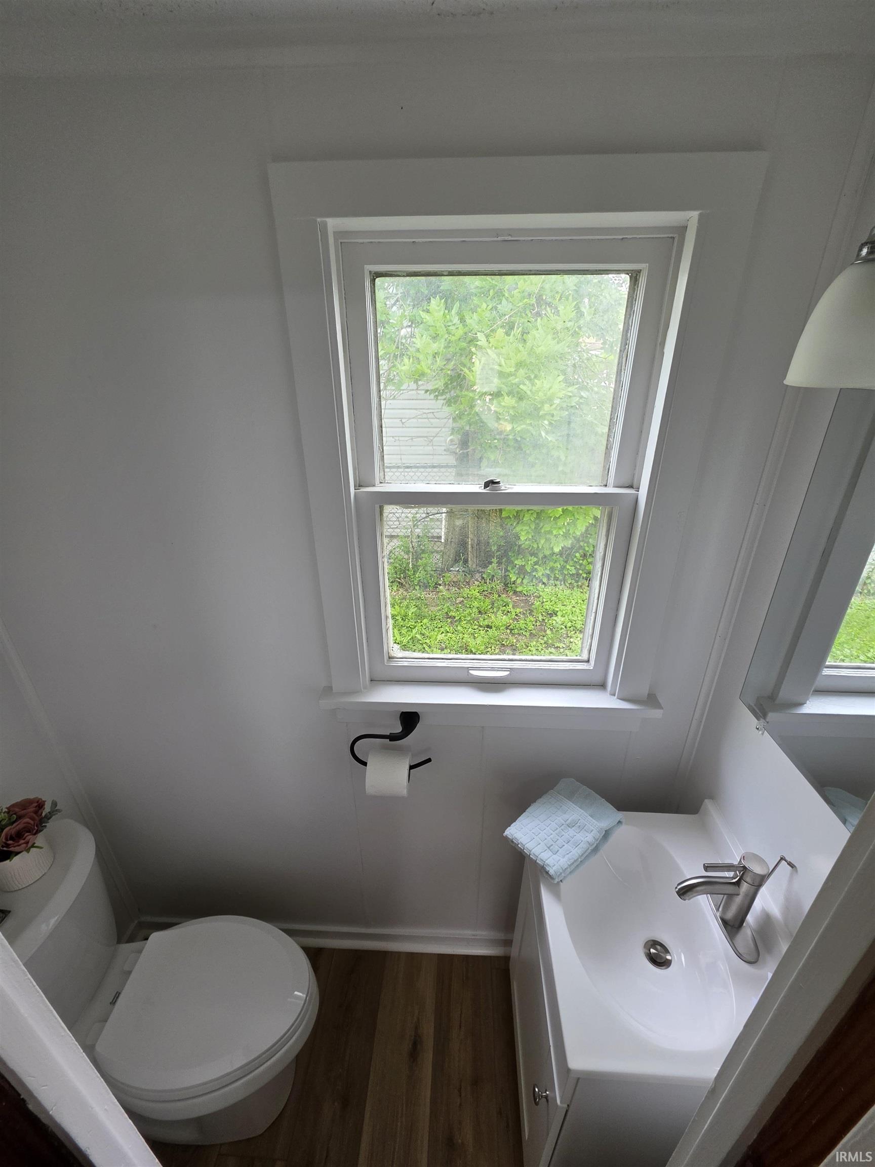 Compact bathroom featuring wood-finish flooring, a white vanity with an integrated sink, a contemporary chrome faucet, a white toilet, and a white-framed window