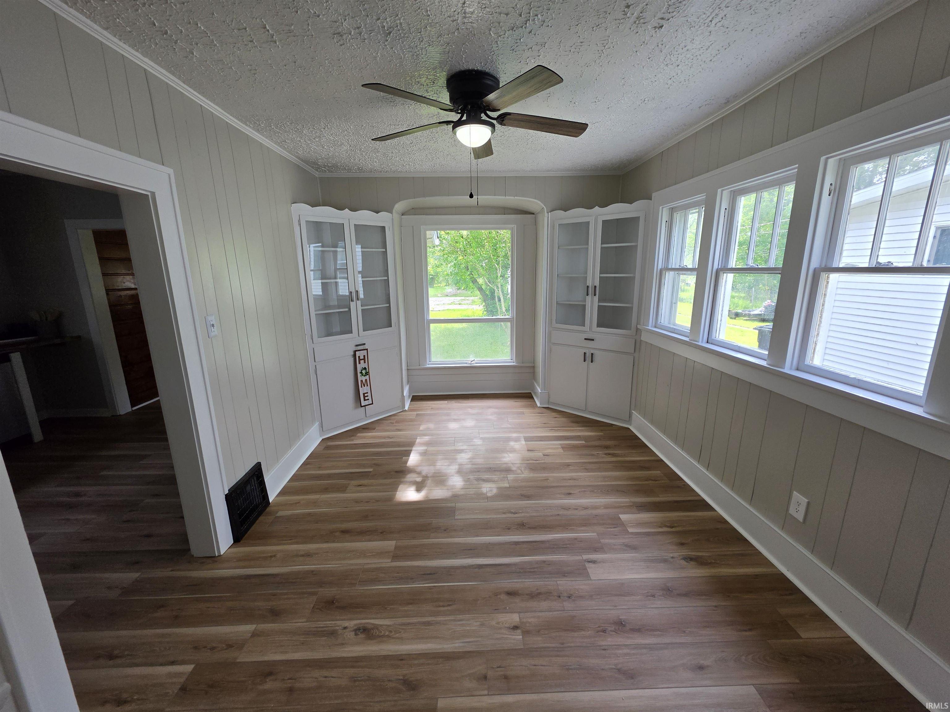 Spacious room with wood-finish flooring, light-toned vertical paneling, white trim, and a ceiling fan with integrated lighting