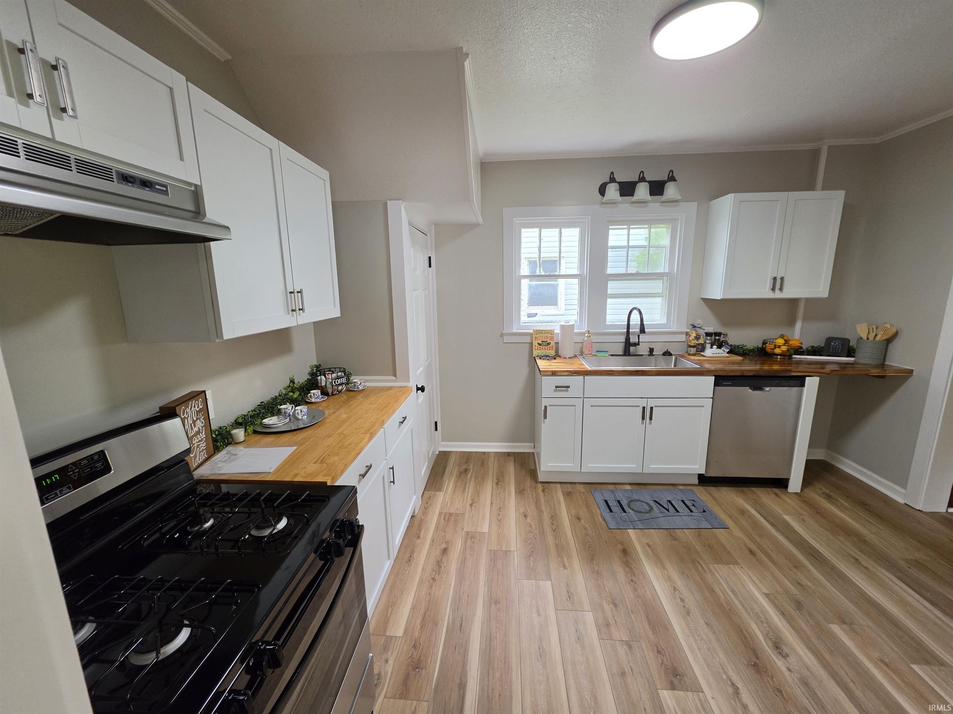Modern kitchen featuring wood-finish flooring, white cabinetry with contemporary hardware, stainless steel appliances, butcher block countertops, and a stainless steel sink with a black-finish faucet