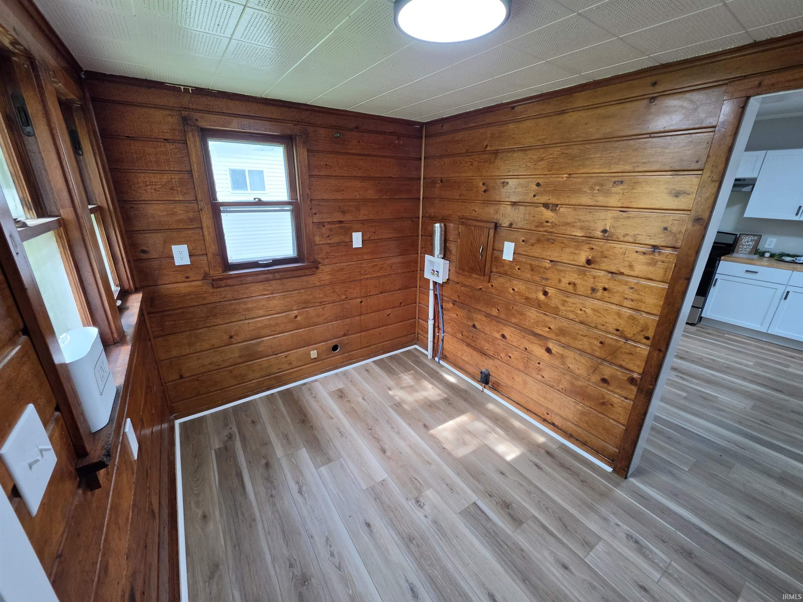 Room featuring wood-finish wall paneling, light wood-finish flooring, a single window with blinds, and white ceiling panels