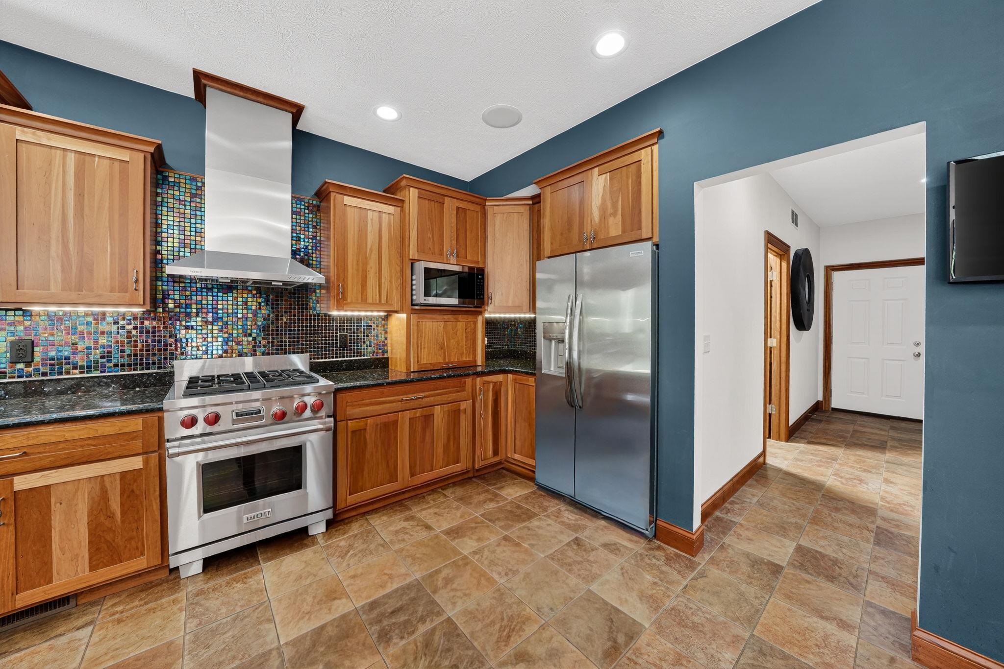 Gourmet kitchen featuring stainless steel appliances, a professional-grade range with a stainless steel vent hood, wood cabinetry, dark countertops, and a mosaic tile backsplash