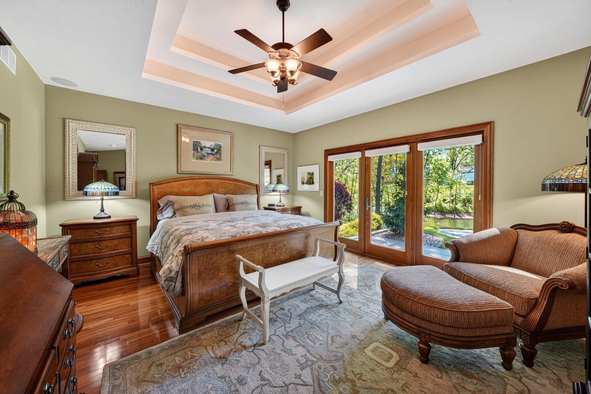 Spacious bedroom featuring warm wood-finish flooring, a tray ceiling with a built-in ceiling fan, and direct access to an outdoor area through a three-panel glass door with wood framing