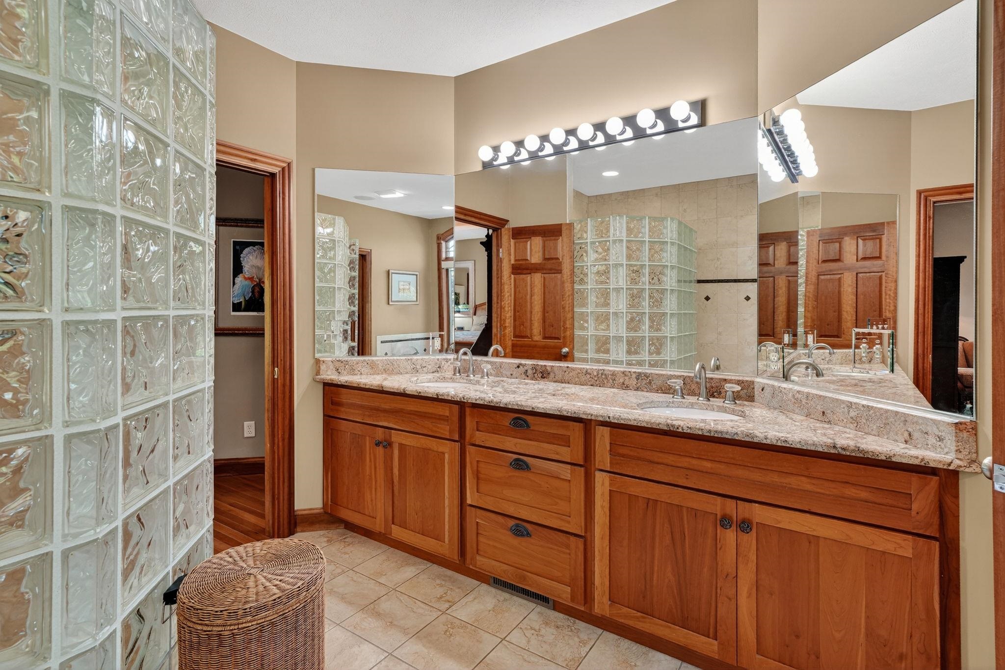 Bathroom featuring a curved glass block partition, double vanity with stone countertop, natural wood cabinetry, and extensive mirror