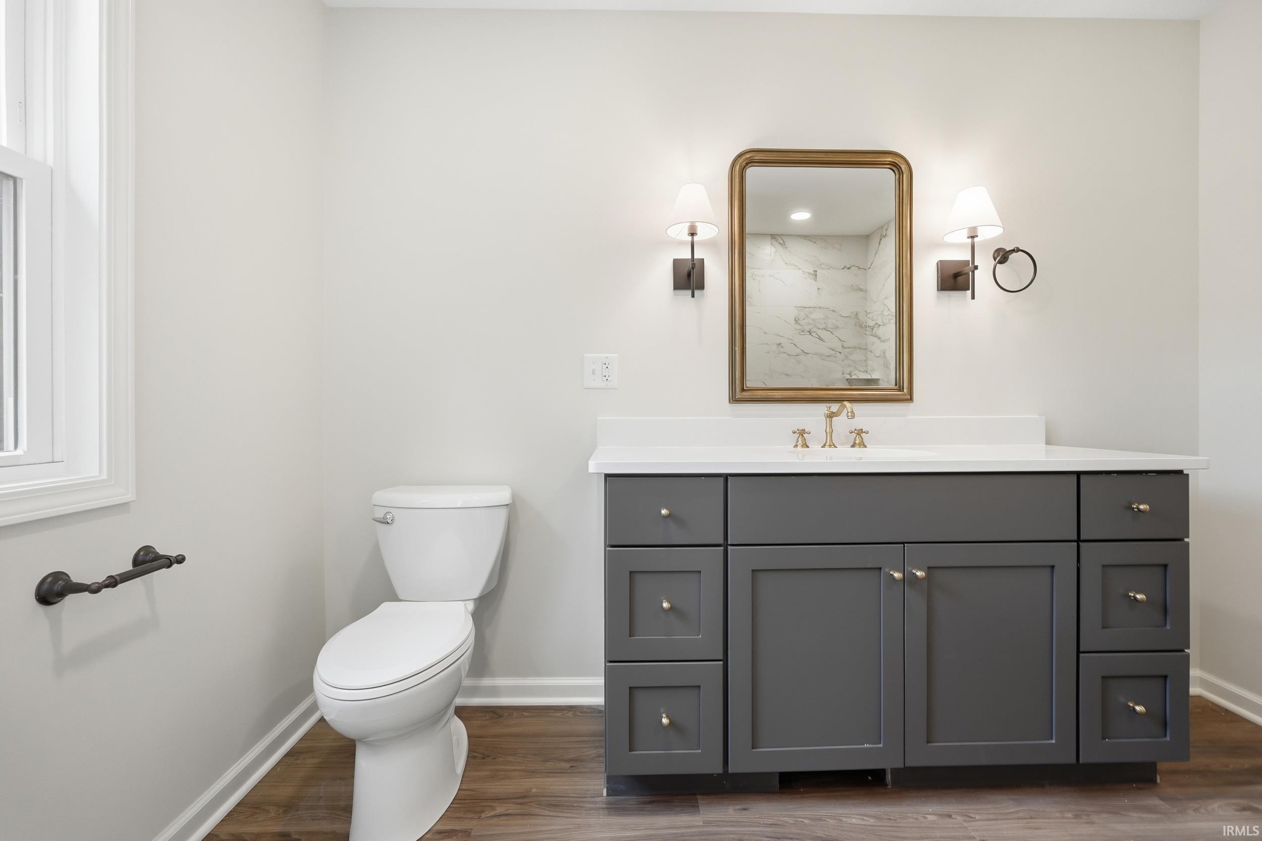 Spacious bathroom featuring a dark grey vanity with a white countertop, gold-finish fixtures, dual wall sconces, and a rectangular mirror with a gold-tone frame