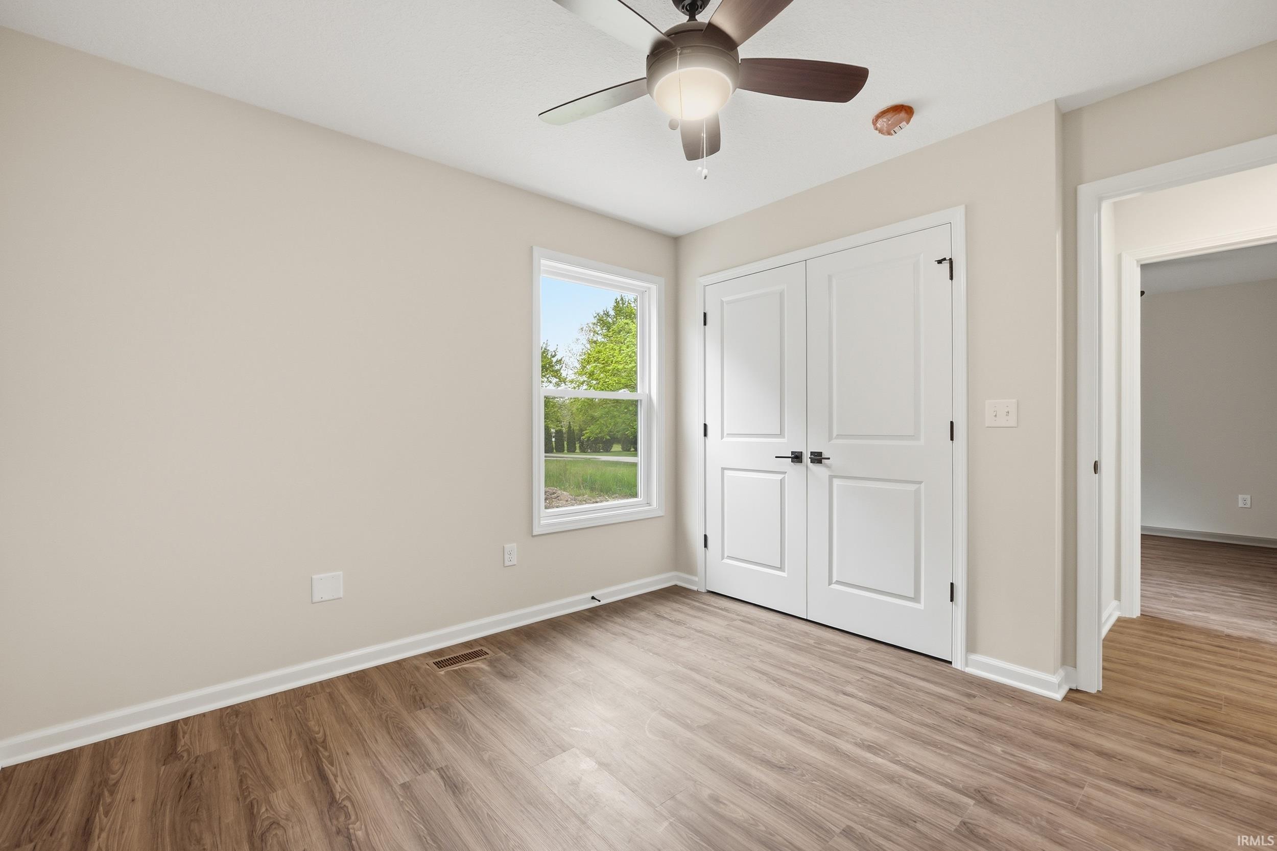 Neutral-toned interior featuring wood-finish flooring, a ceiling fan with integrated lighting, a double-door closet with dark hardware, and a window with white trim