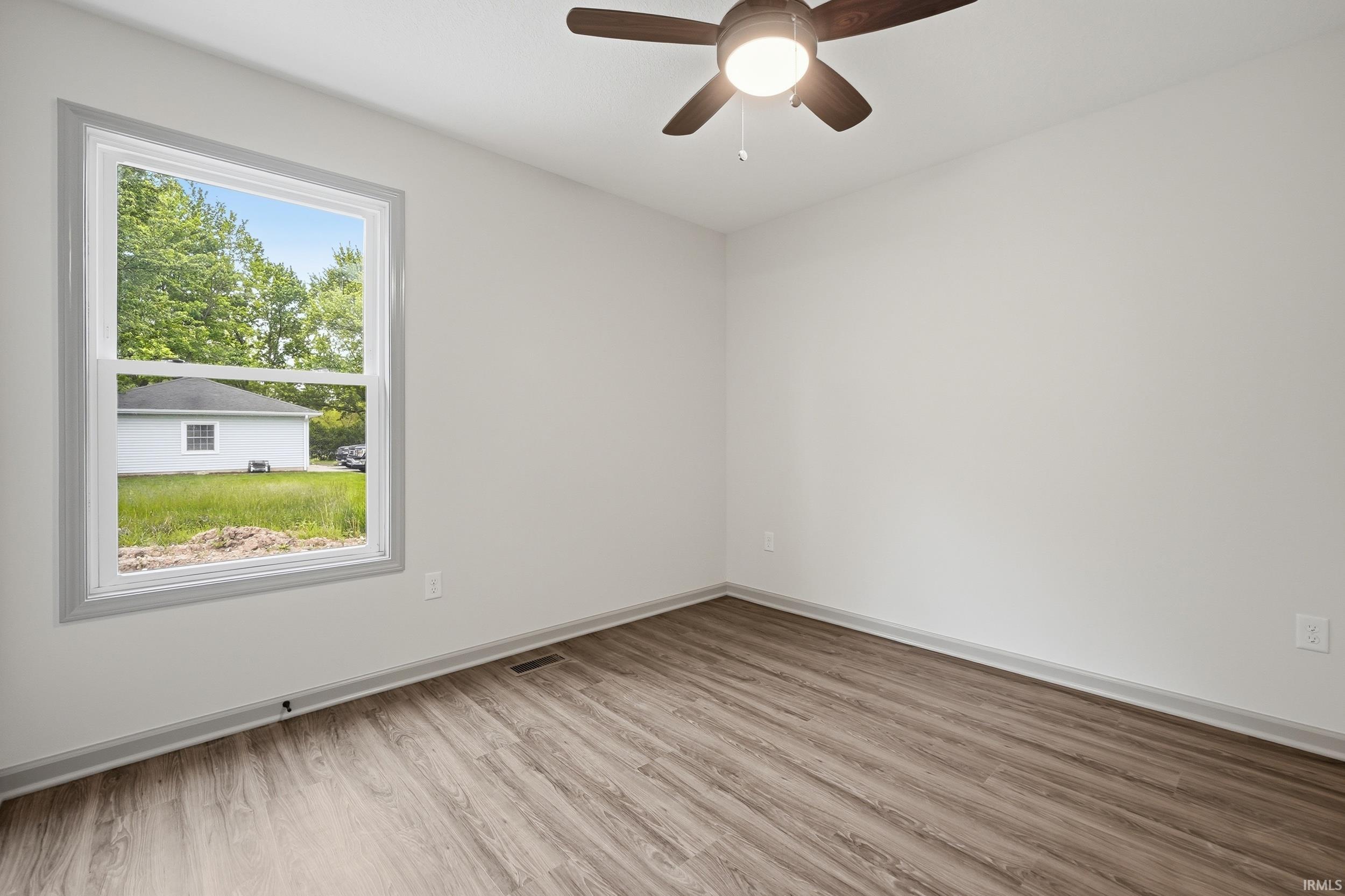 Bright interior room featuring wood-finish flooring, a ceiling fan with integrated lighting, white walls, and a large window with gray trim