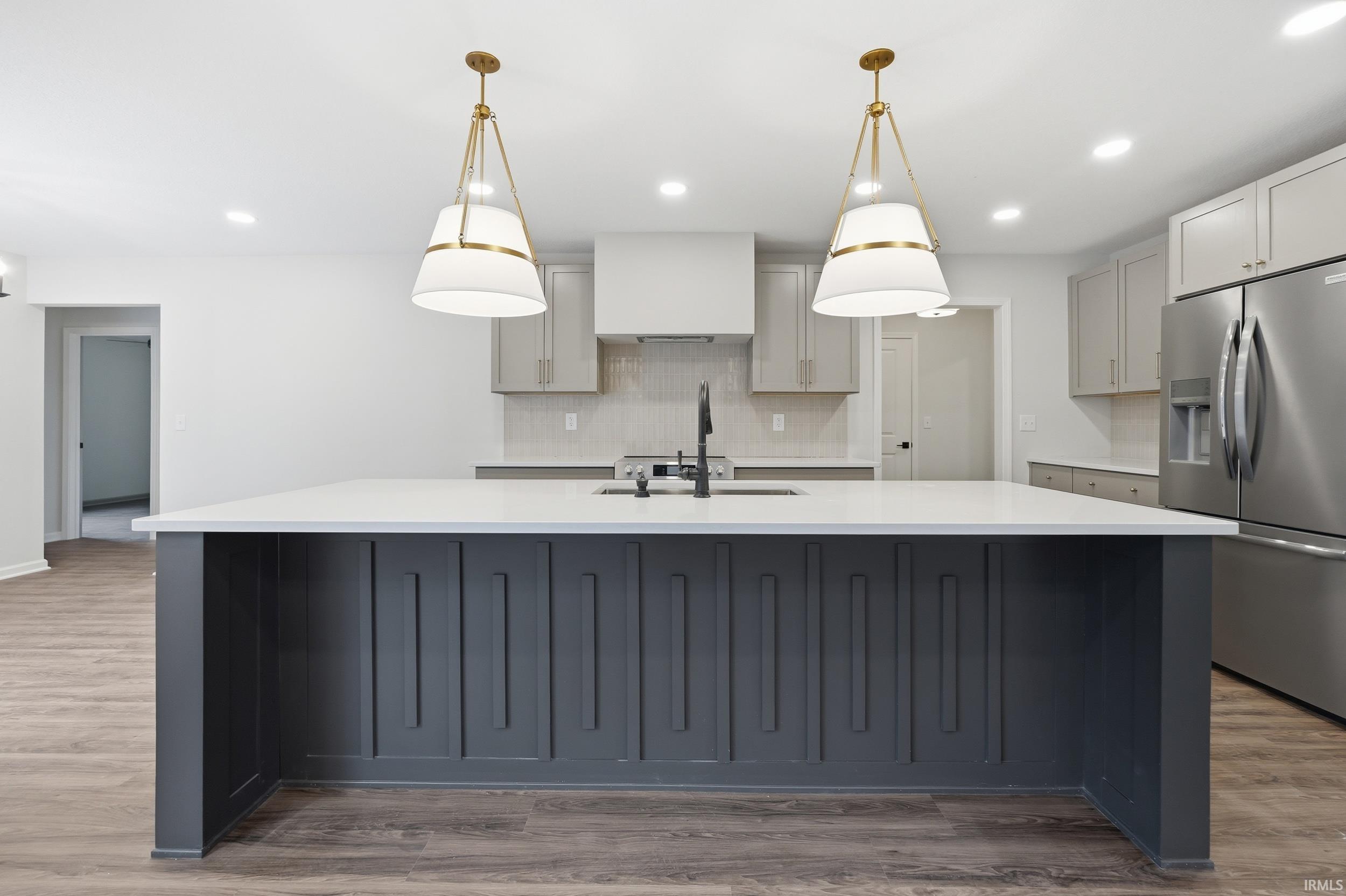 Kitchen featuring a large central island with a white countertop and dark gray paneling, two brass and white pendant lights, light gray cabinetry, stainless steel refrigerator, and wood-finish flooring