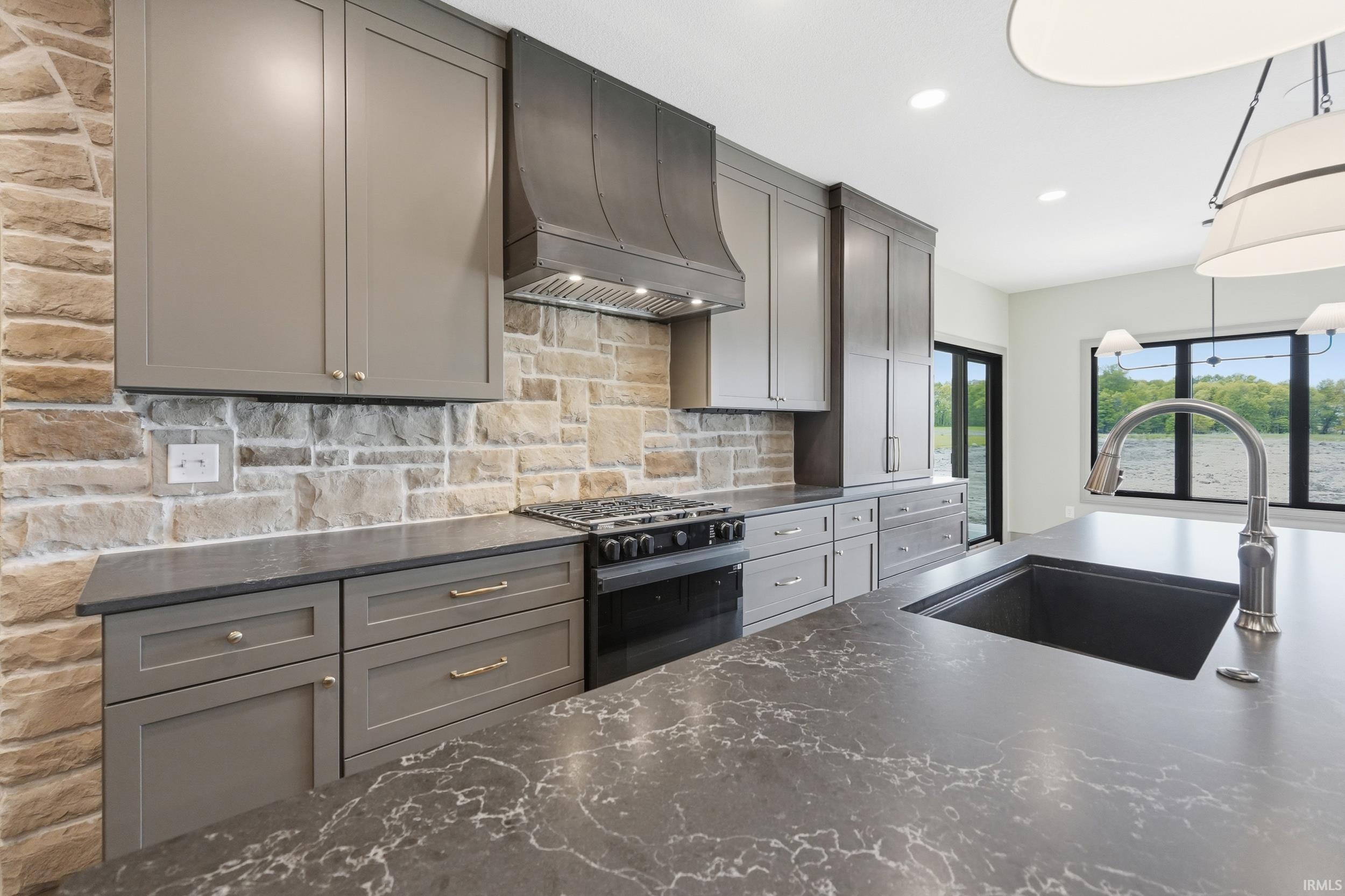 Gourmet kitchen featuring a stone accent wall, custom cabinetry, dark quartz countertops, a gas range with custom hood, and an oversized island with an undermount sink and gooseneck faucet