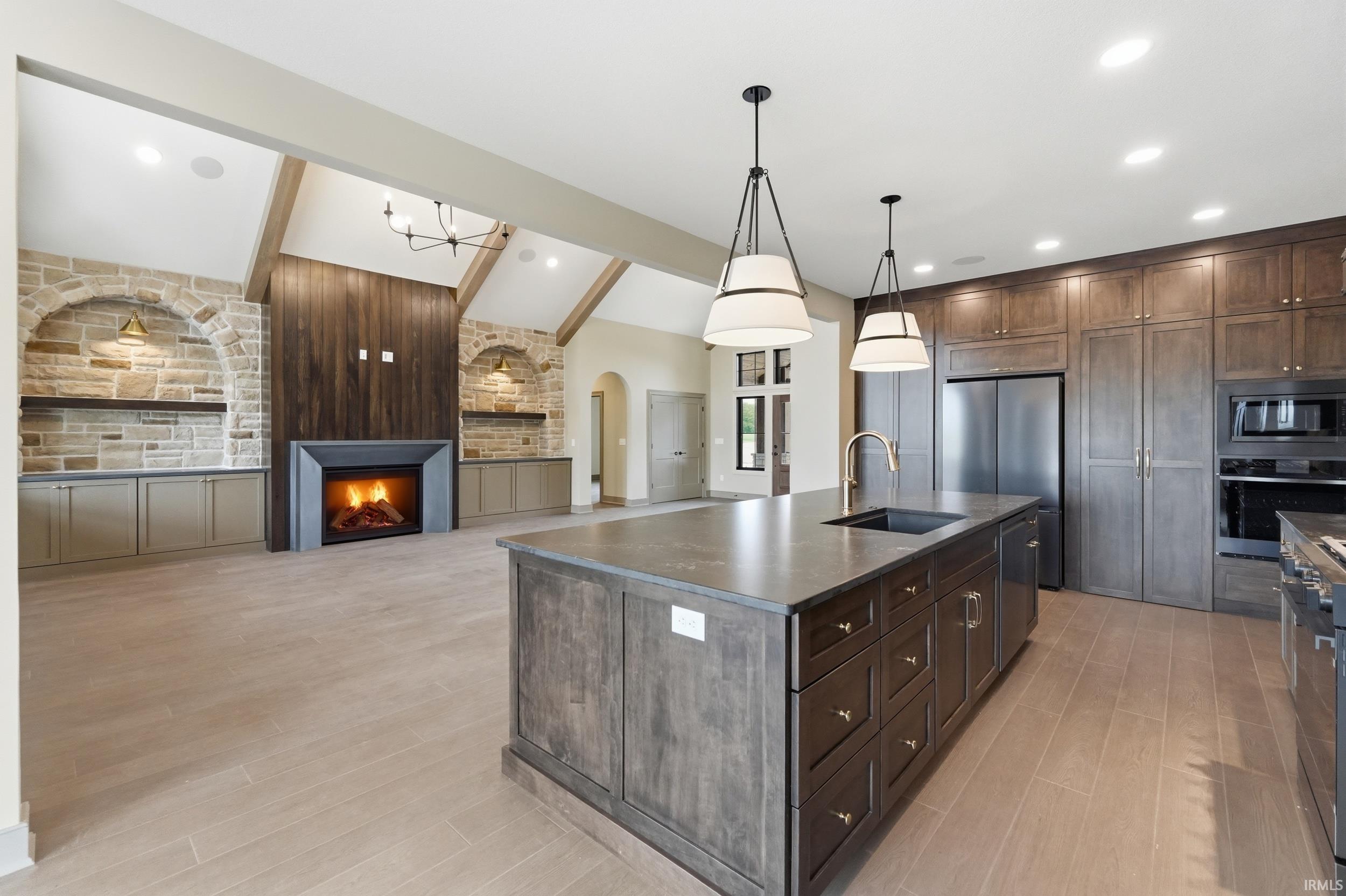 Open-concept living space with vaulted ceilings featuring exposed beams, a stone fireplace with a dark wood-paneled surround, an oversized kitchen island with a built-in sink, stainless steel appliances, and wood-finish flooring