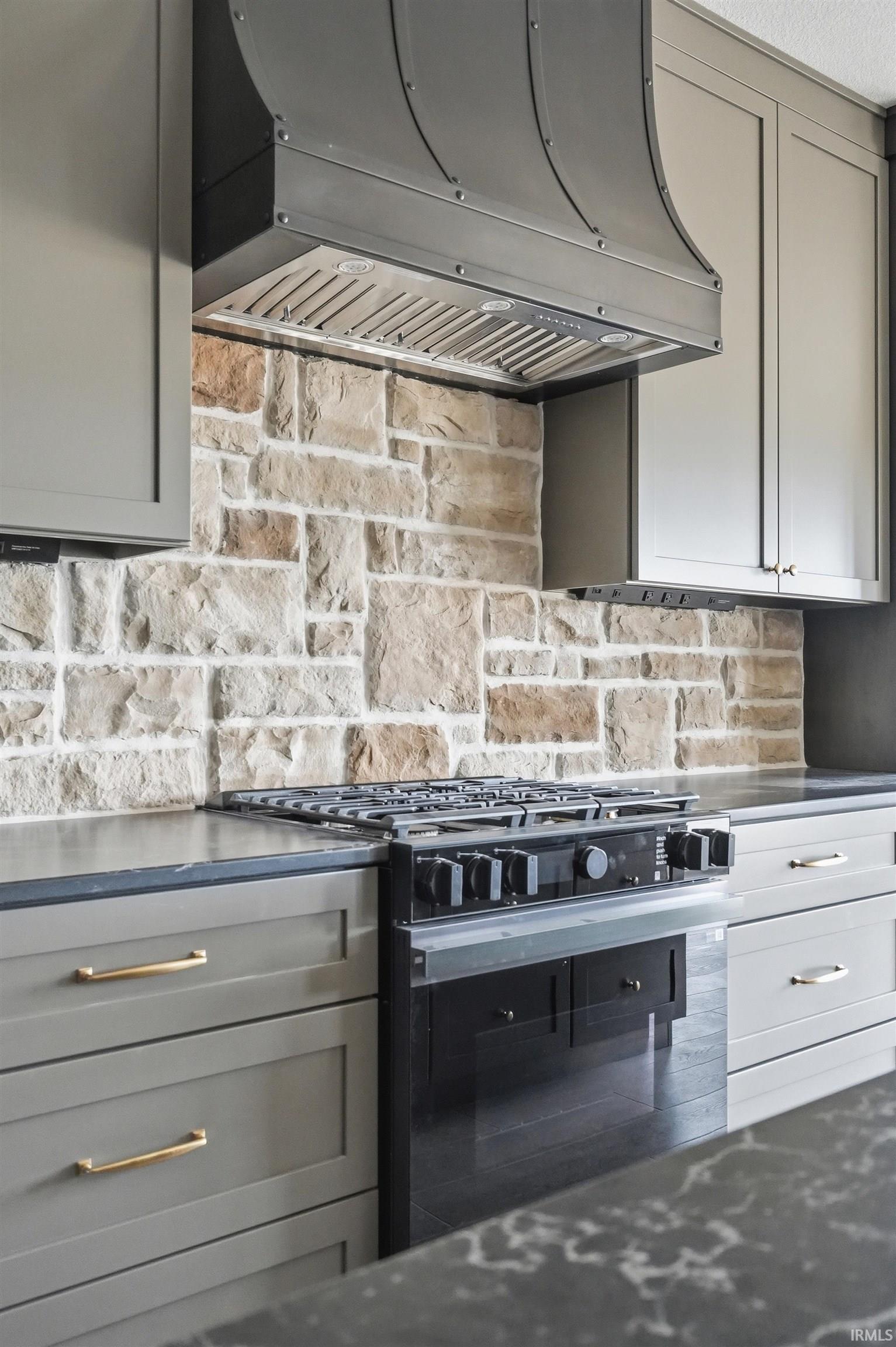 Gourmet kitchen featuring a stone block backsplash, custom range hood, gas range, dark cabinetry, and gold-tone hardware