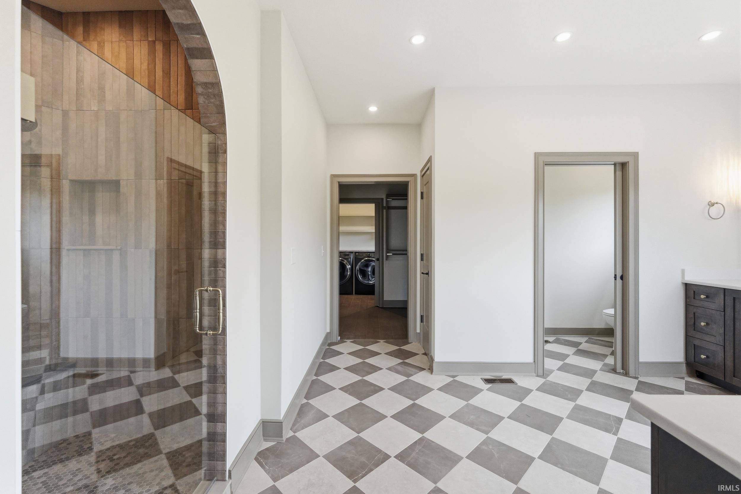 Spacious bathroom featuring checkered tile flooring, a glass-enclosed shower with an arched wood-finish ceiling, recessed lighting, a dark wood-finish vanity with a light countertop, and multiple doorways leading to additional areas