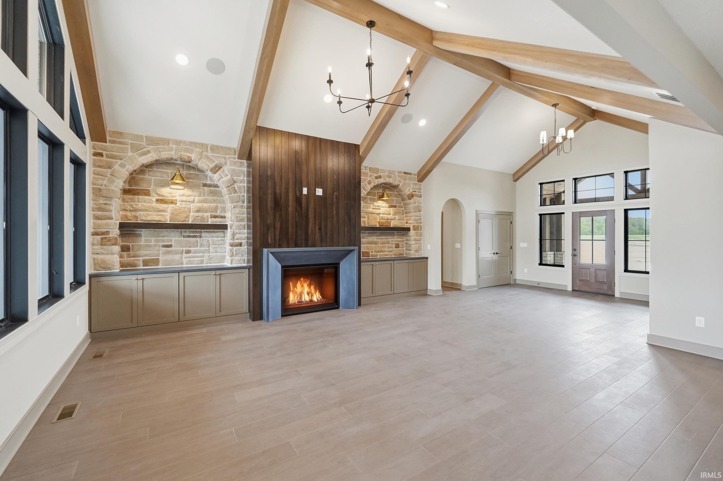 Spacious living area featuring a vaulted ceiling with exposed wood beams, a stone fireplace with a dark wood accent wall, built-in cabinetry with arched stone alcoves, and light-colored flooring