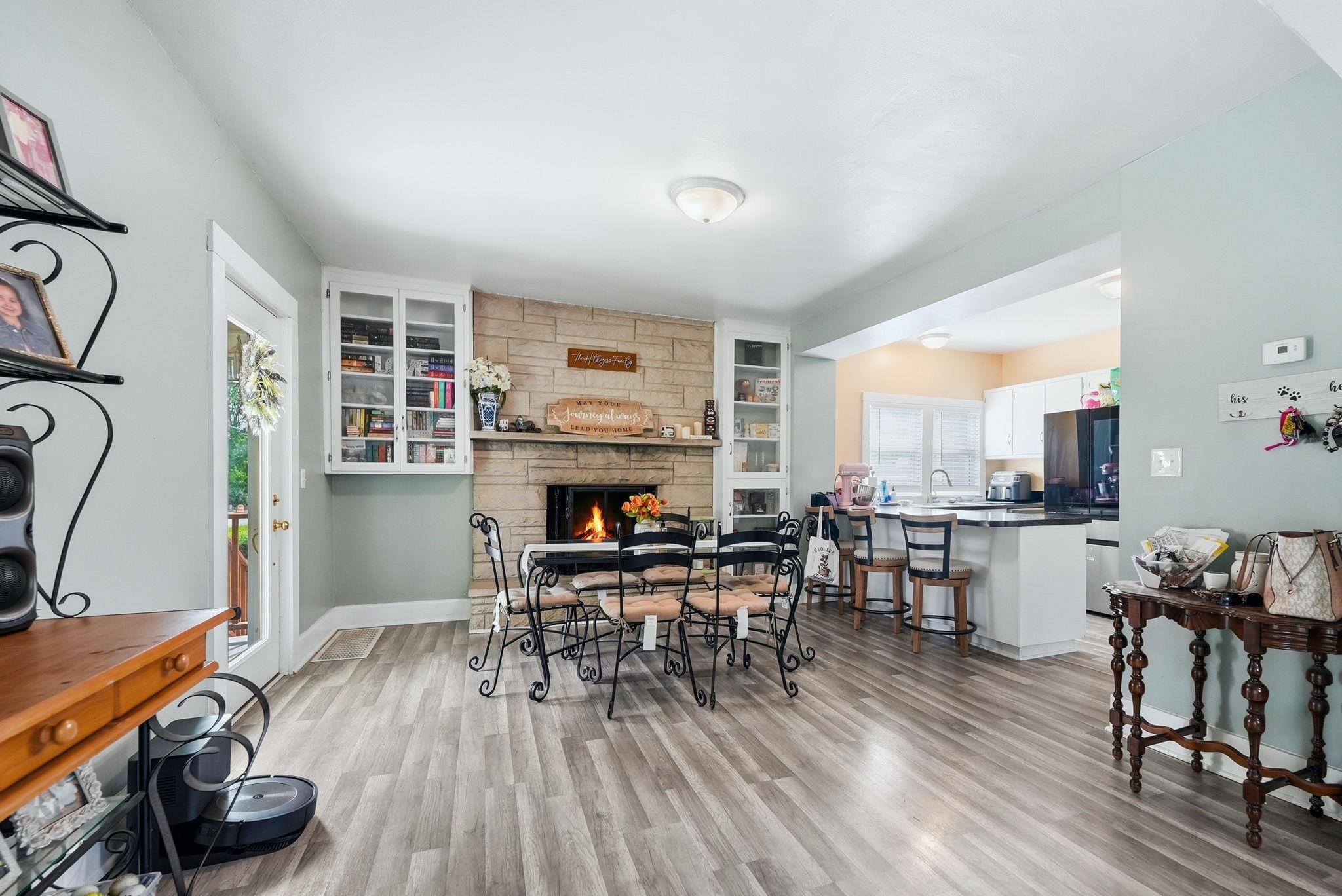Spacious room featuring wood-finish flooring, a stone-clad fireplace with built-in cabinetry, and an open concept kitchen with white cabinetry and a black countertop