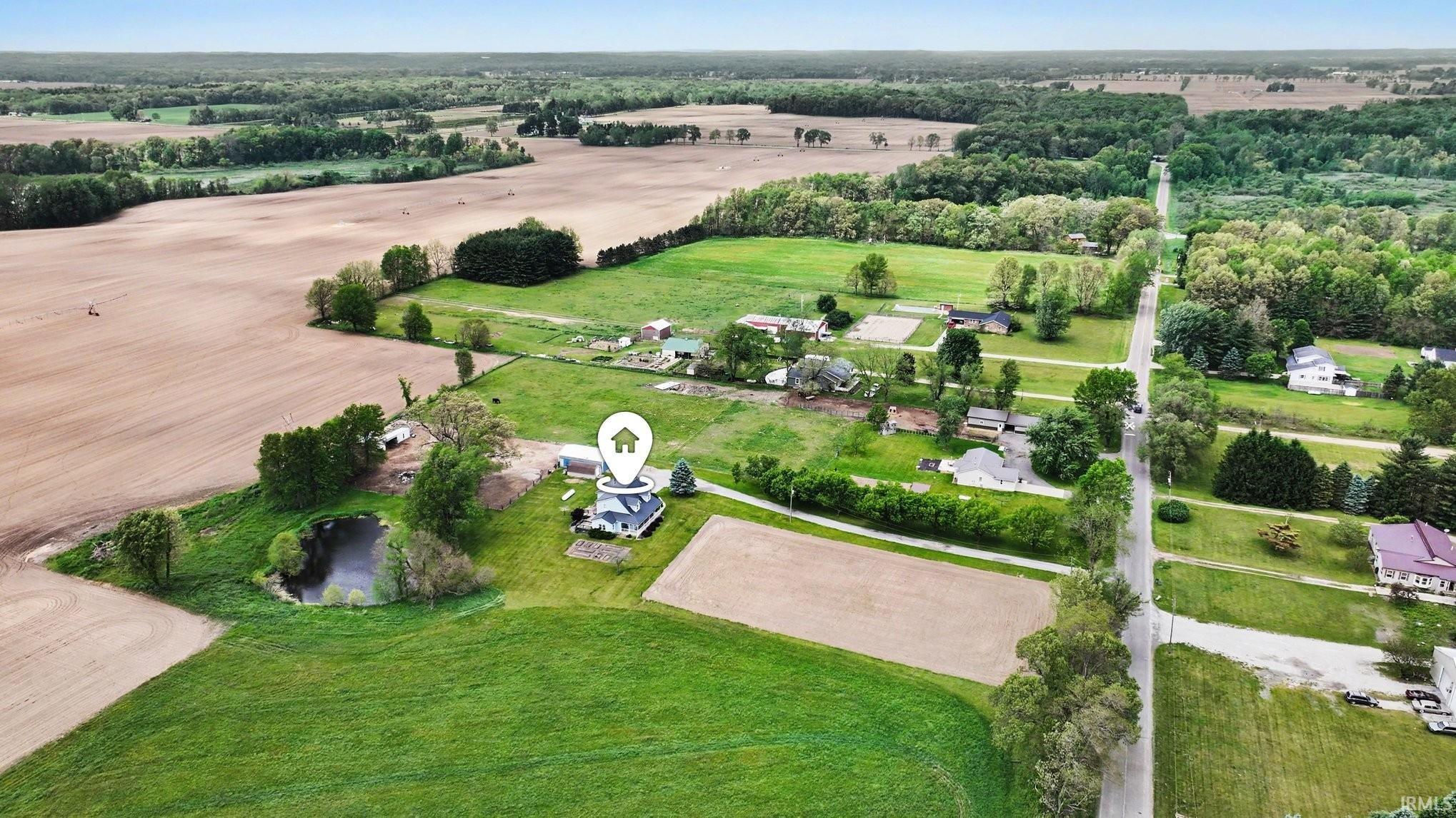 Aerial view showcasing the property with expansive green space, a private pond feature, a mature tree line, and paved road frontage