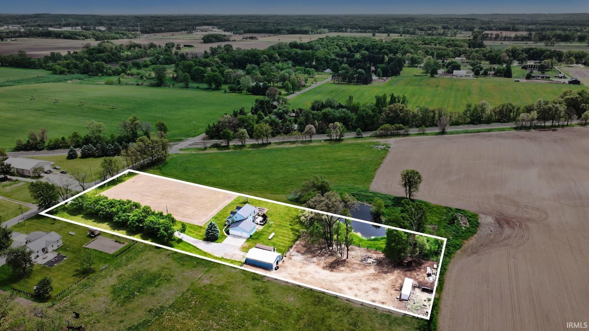 Expansive rural property featuring a primary residence with a gray roof and light blue siding, a detached garage, a large cultivated field, a natural pond, and mature trees outlining the property