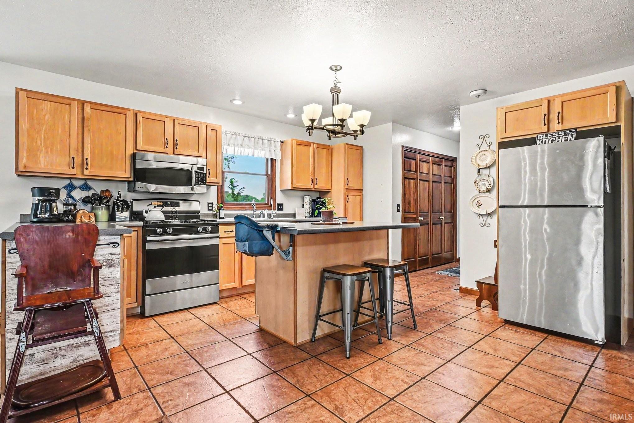 Kitchen featuring wood-finish cabinetry, stainless steel appliances, a center island with seating, pendant lighting, and terracotta-toned tile flooring