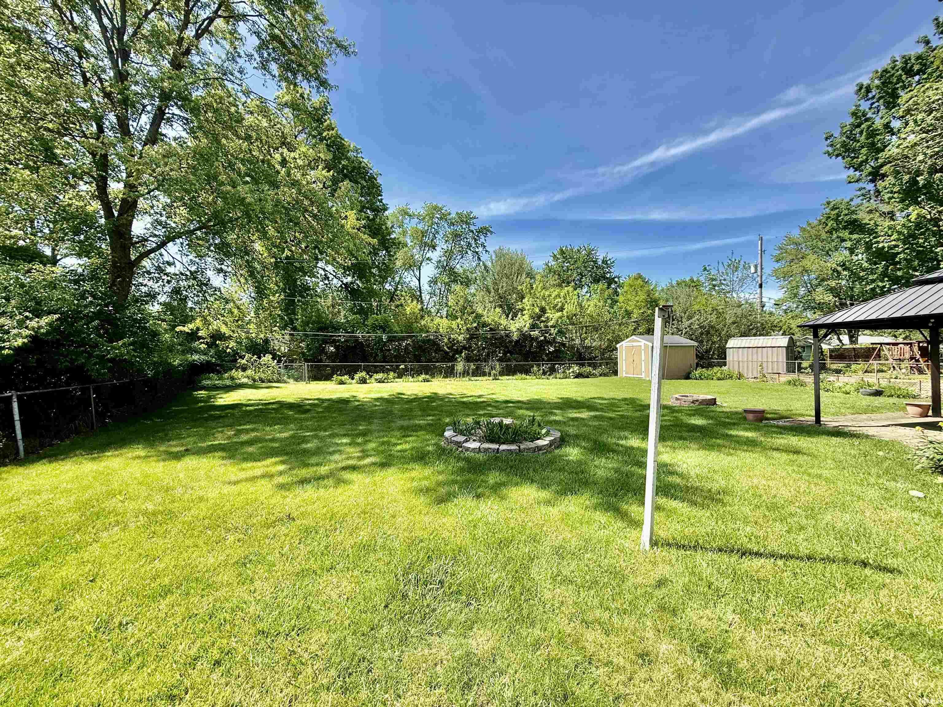 Expansive grassy yard featuring a chain-link fence, mature trees, two storage sheds, and a permanent gazebo structure