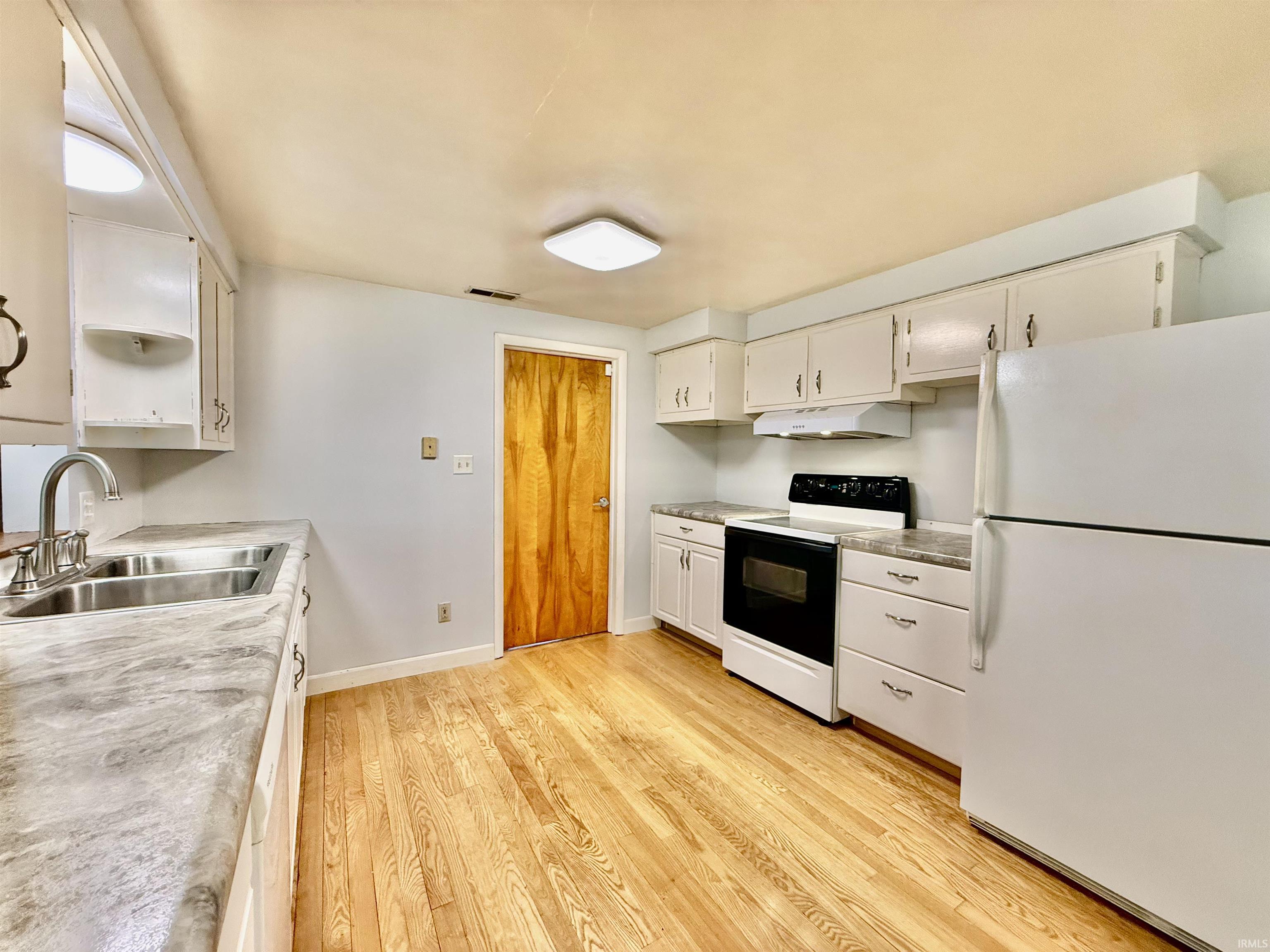 Kitchen featuring wood-finish flooring, white cabinetry with silver hardware, dual basin stainless steel sink with a gooseneck faucet, white appliances, and a wood-panel door