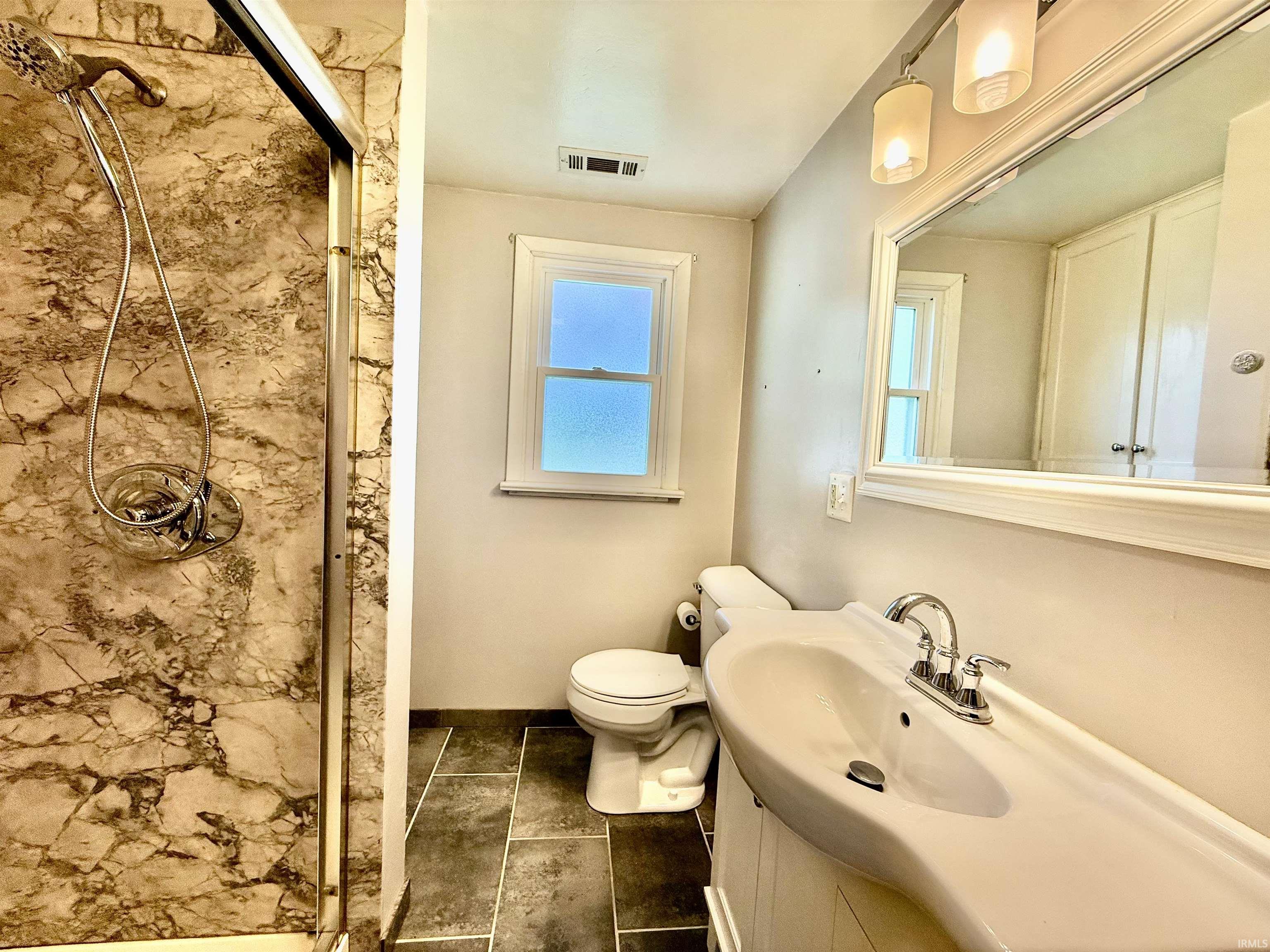 Bathroom featuring a walk-in shower with stone-patterned walls, dark tile flooring, a single window, and a vanity with an integrated sink