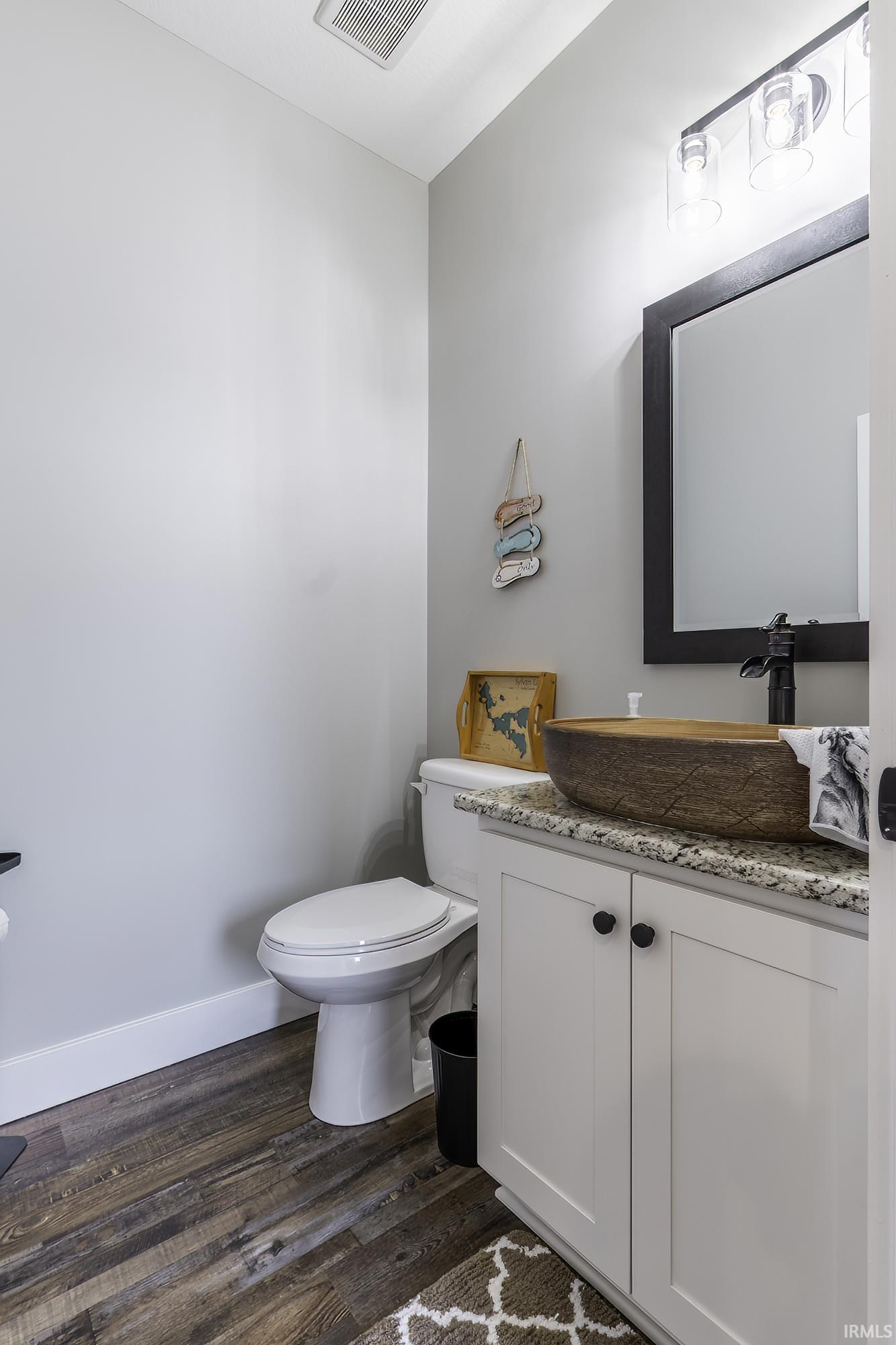 Powder room featuring a unique wood-finish vessel sink, white shaker-style vanity with dark hardware, granite countertop, and wood-finish flooring