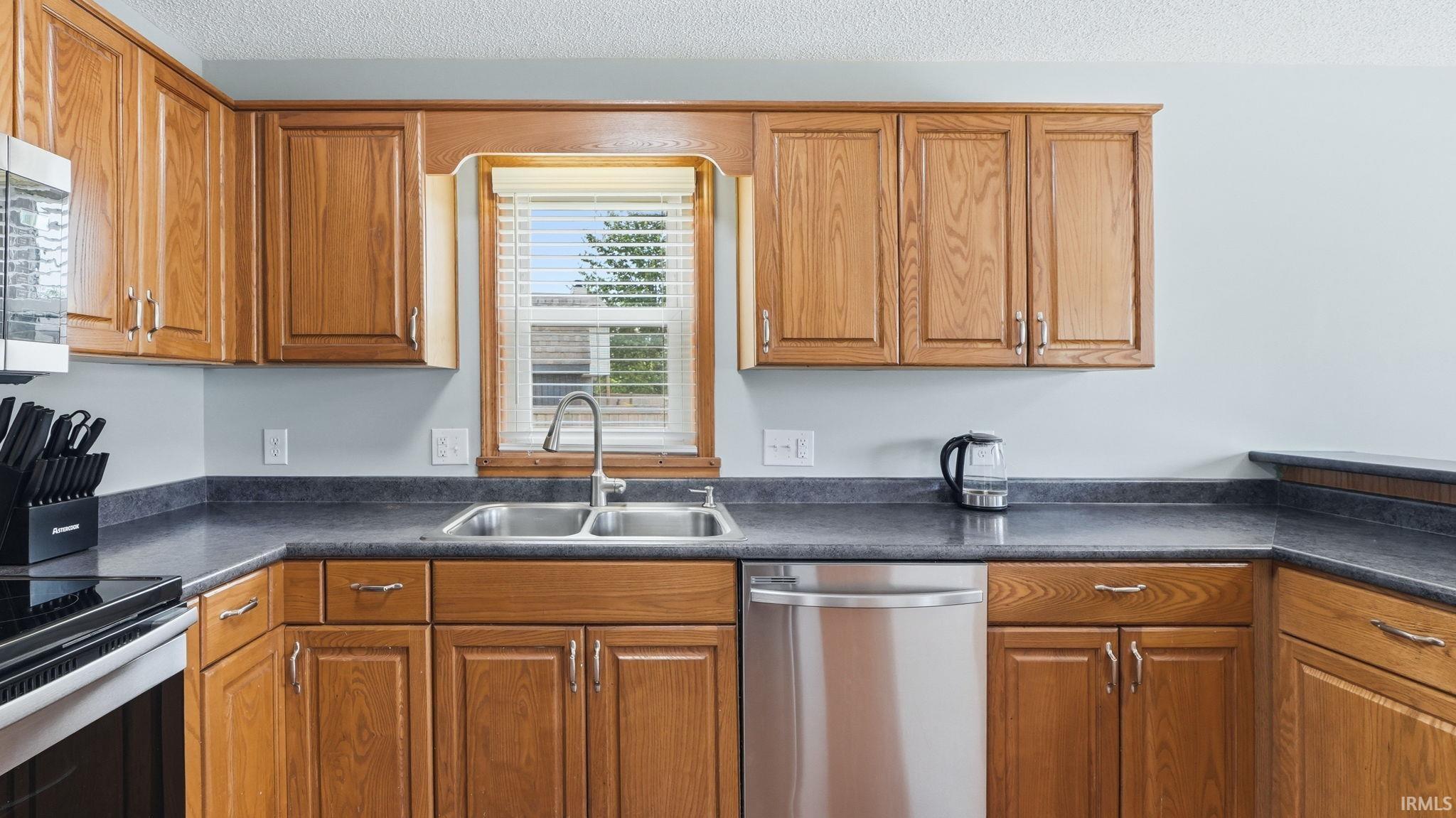 Kitchen featuring wood-finish cabinetry, dark countertops, a double basin stainless steel sink, and a stainless steel dishwasher