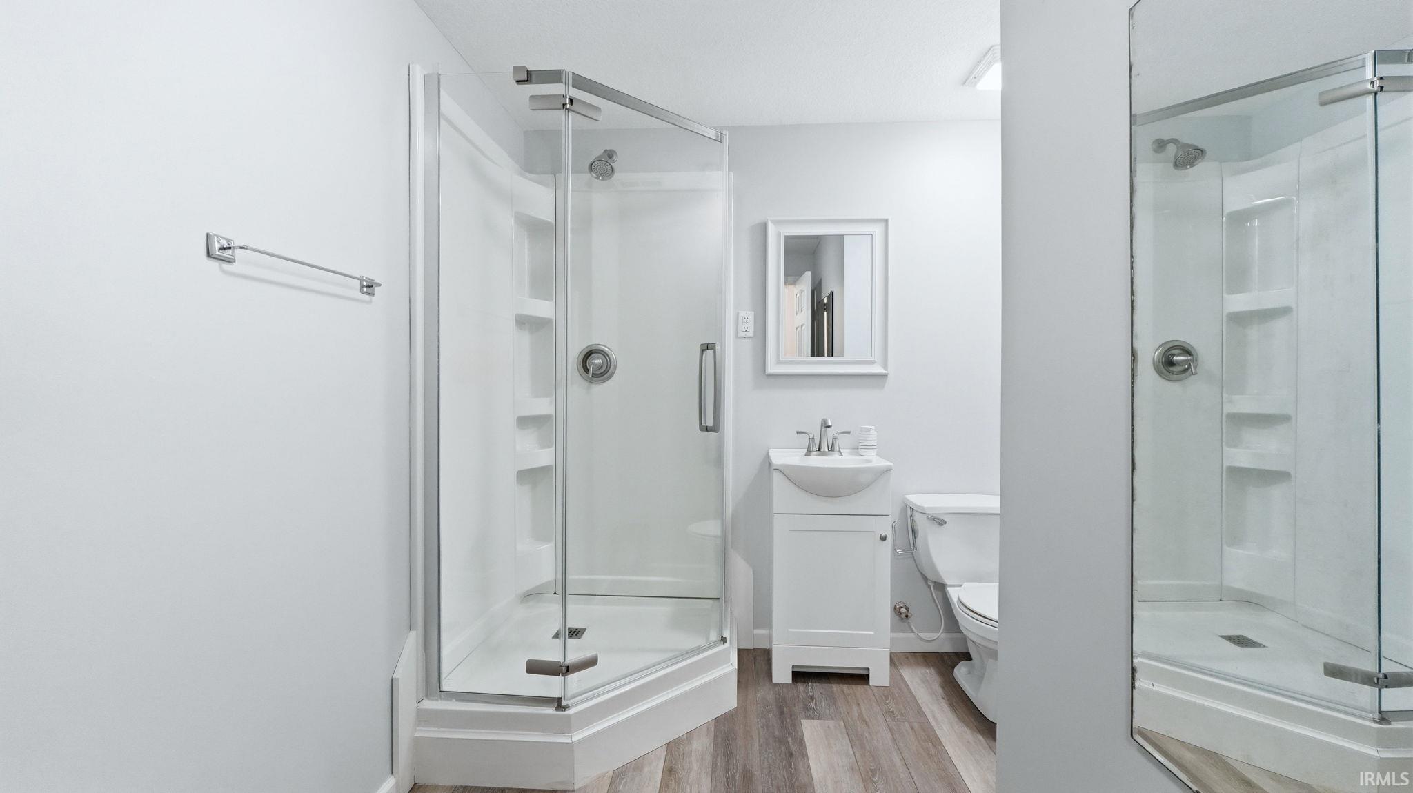Corner glass-enclosed shower with integrated shelving, a white pedestal sink with vanity storage, wood-finish flooring, and a wall-mounted mirror