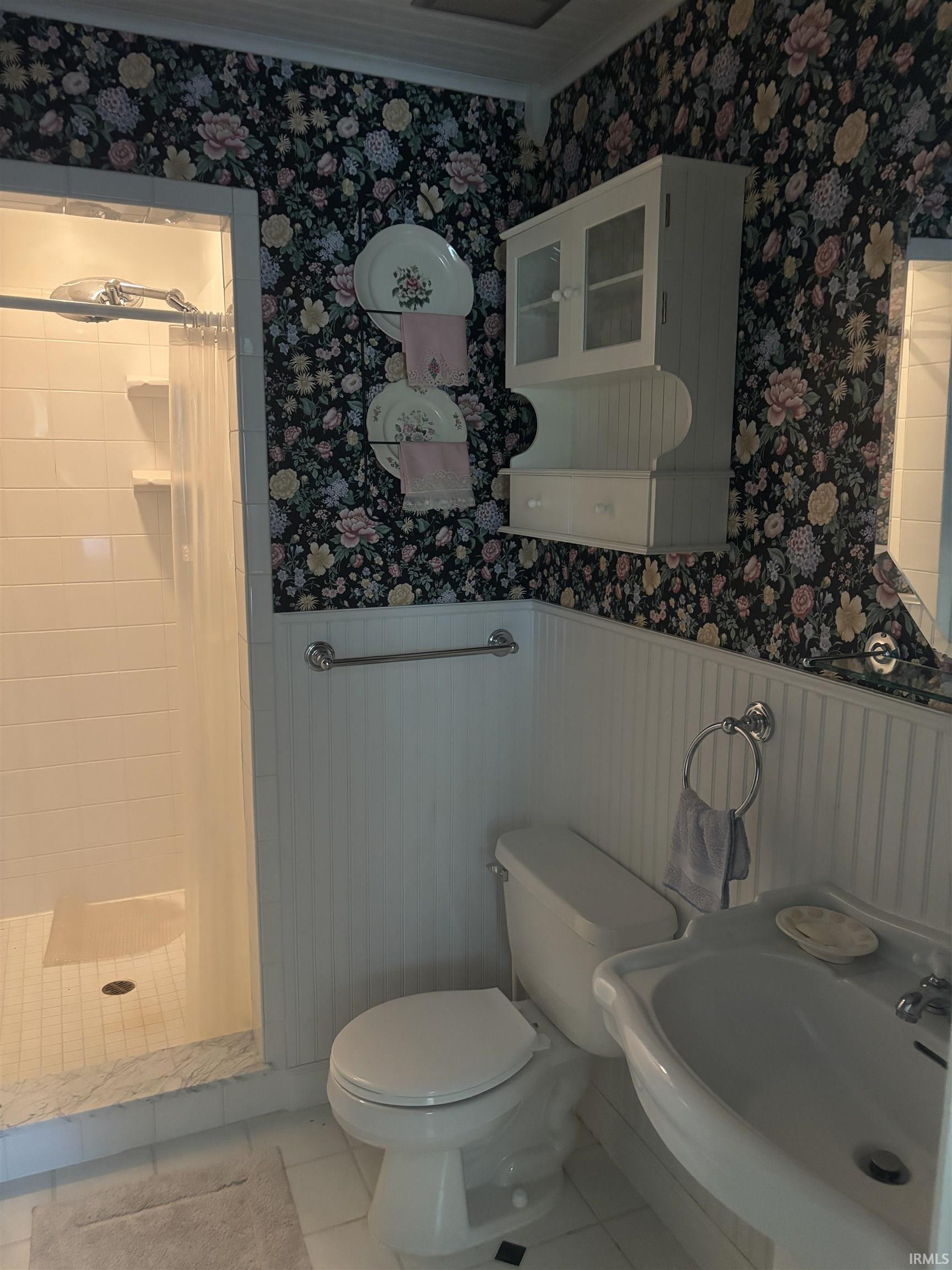 Tiled shower enclosure with built-in shelving, pedestal sink, wall-mounted storage cabinet, and beadboard paneling