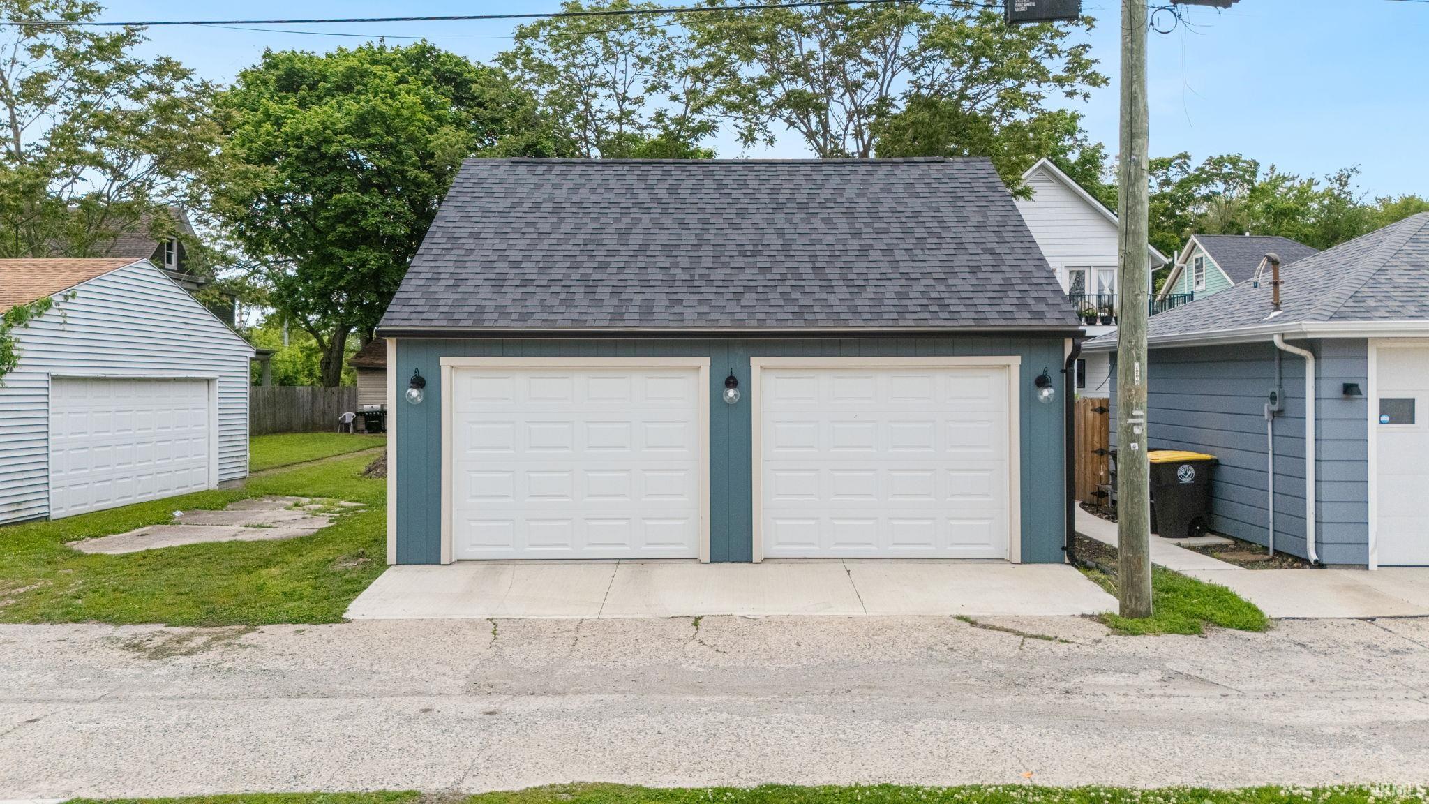 Detached two-car garage featuring a dark shingle roof, blue siding, dual white paneled garage doors, and exterior wall sconces