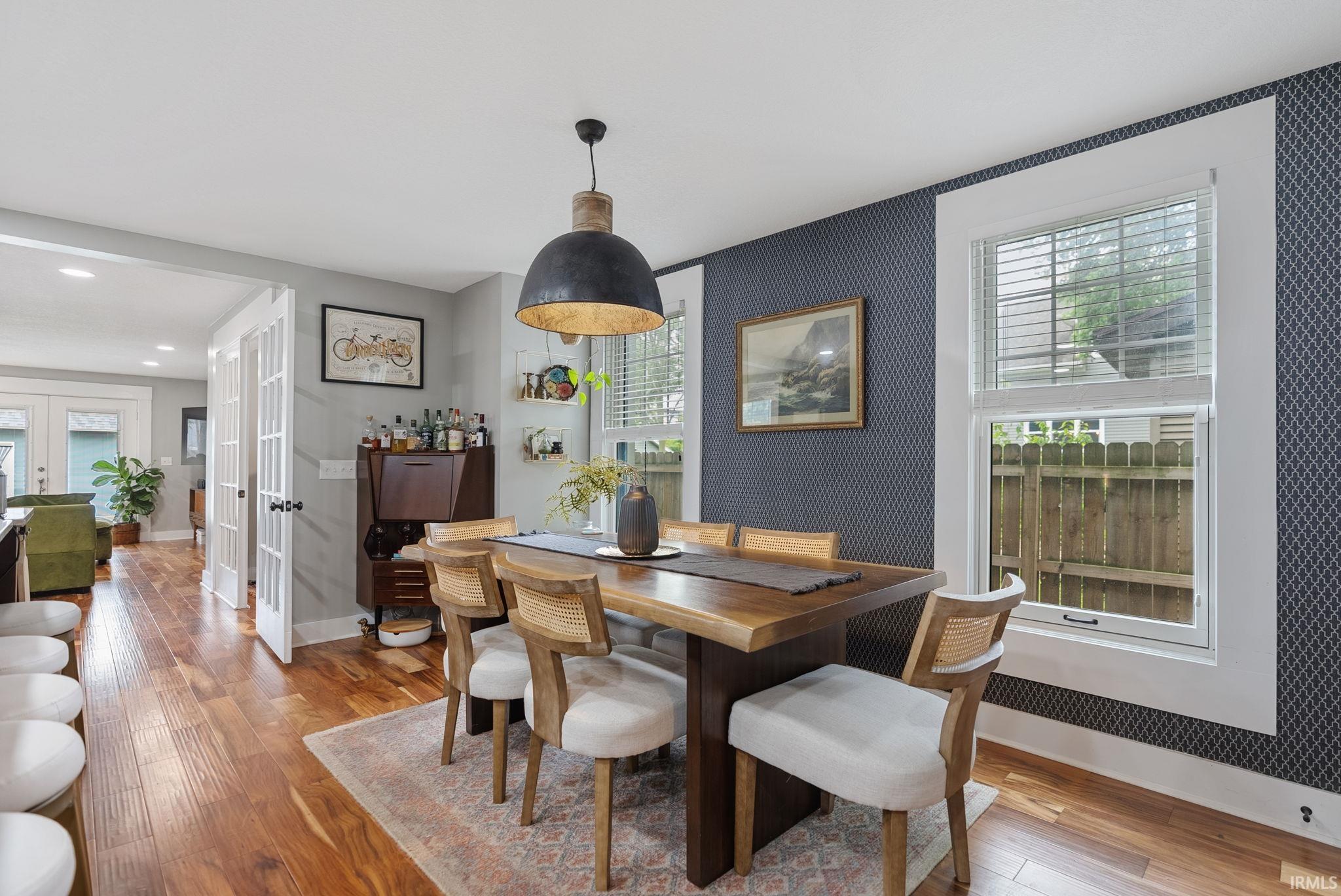 Dining area featuring wood-finish flooring, a contemporary pendant light fixture, patterned accent wall, and white-framed windows