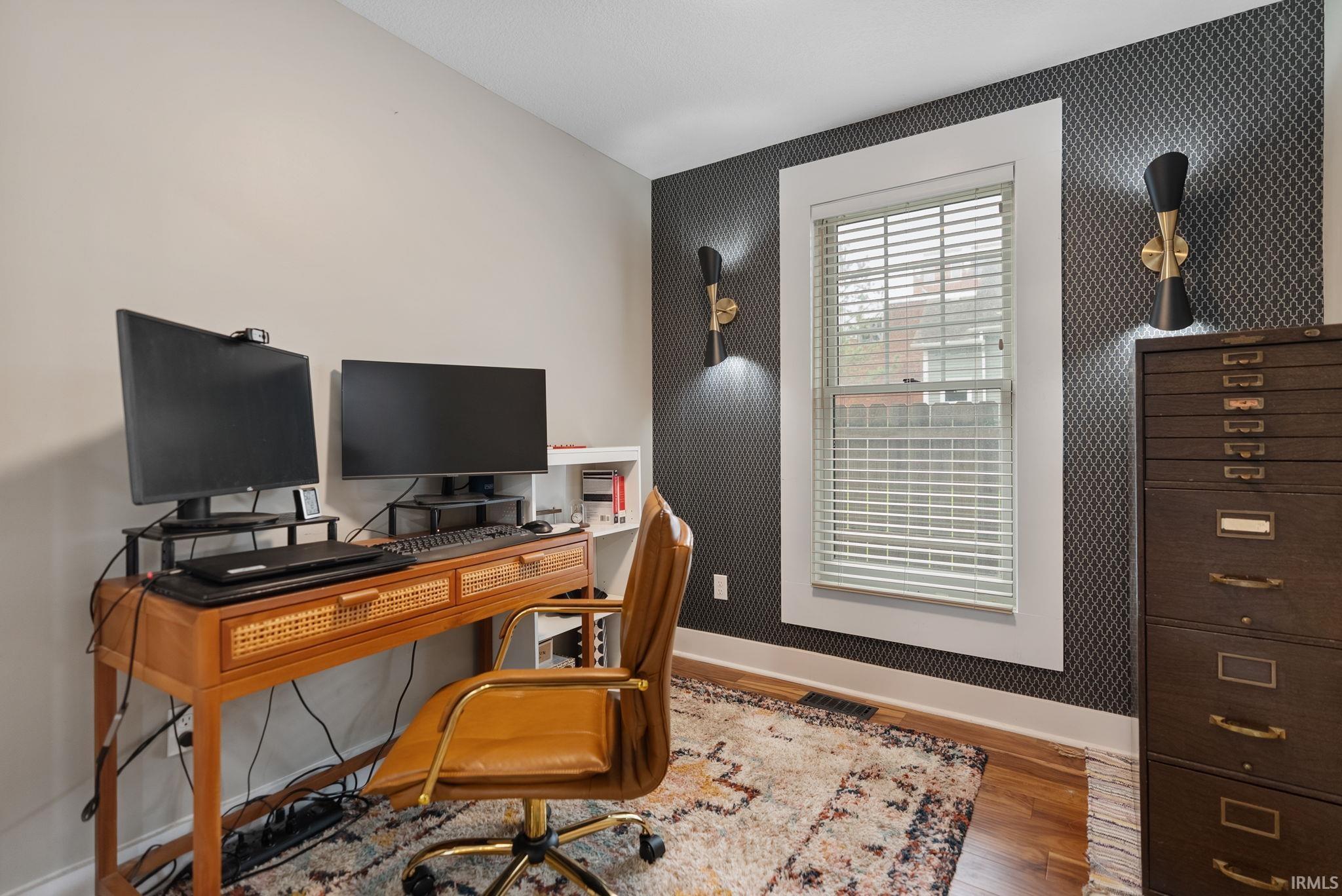 Work area featuring hardwood flooring, a window with white trim and blinds, dark patterned wallpaper, and two wall-mounted sconces
