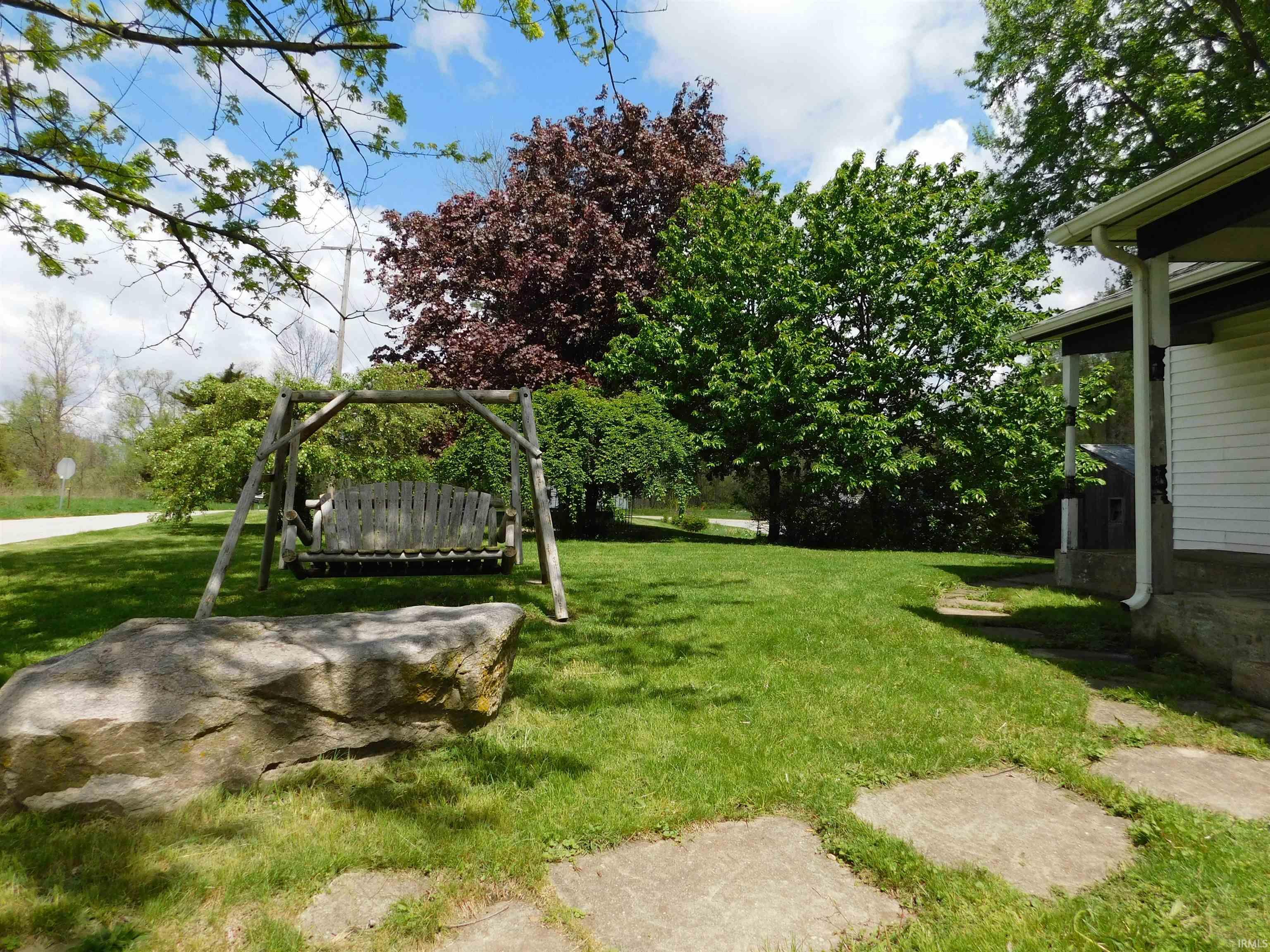 Expansive lawn featuring a wooden porch swing, large natural boulder, mature trees, and concrete stepping stones
