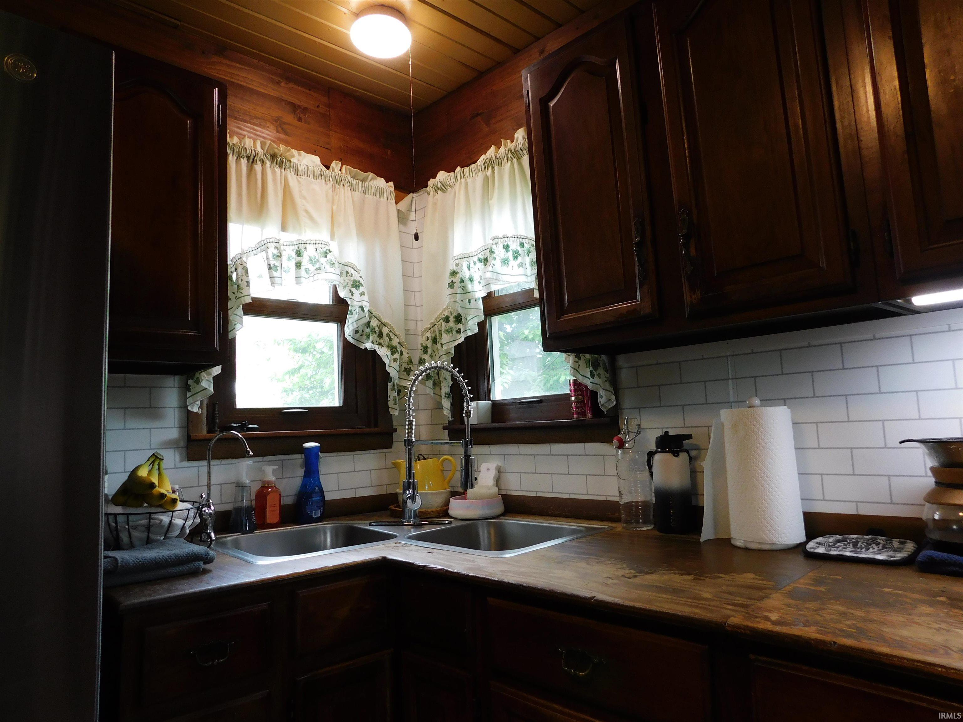 Kitchen featuring a double basin stainless steel sink, wood-finish countertops, subway tile backsplash, dark wood cabinetry, and a wood plank ceiling