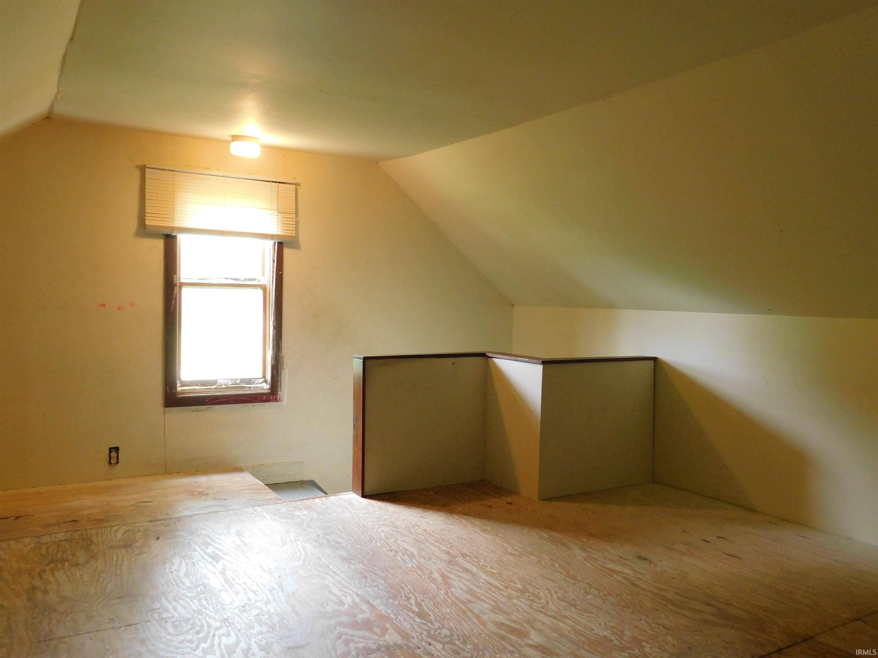 Attic space featuring plywood subflooring, an angled ceiling, a single window with blinds, and built-in knee-wall storage