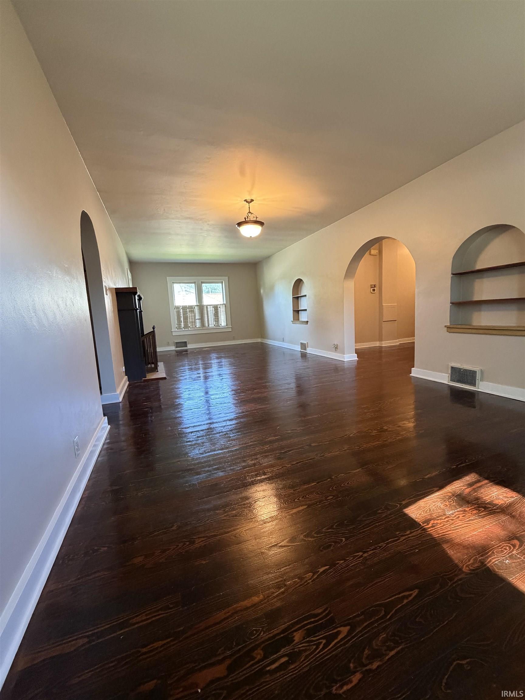 Spacious interior featuring dark wood-finish flooring throughout, multiple architectural archways, built-in wall shelving, and a prominent ceiling-mounted light fixture