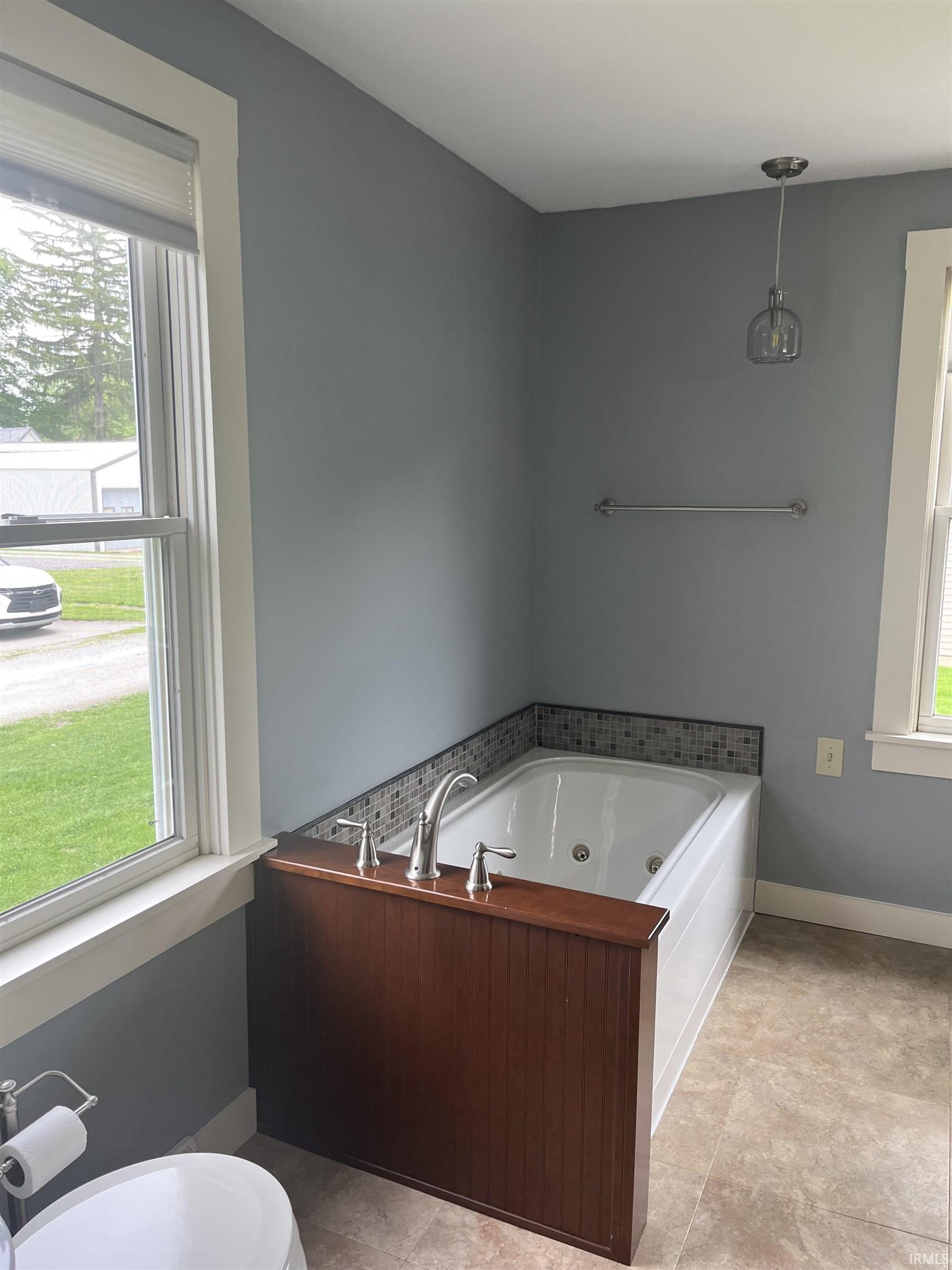 Jetted tub with wood-finish paneling and tile surround, accompanied by a window and pendant light fixture