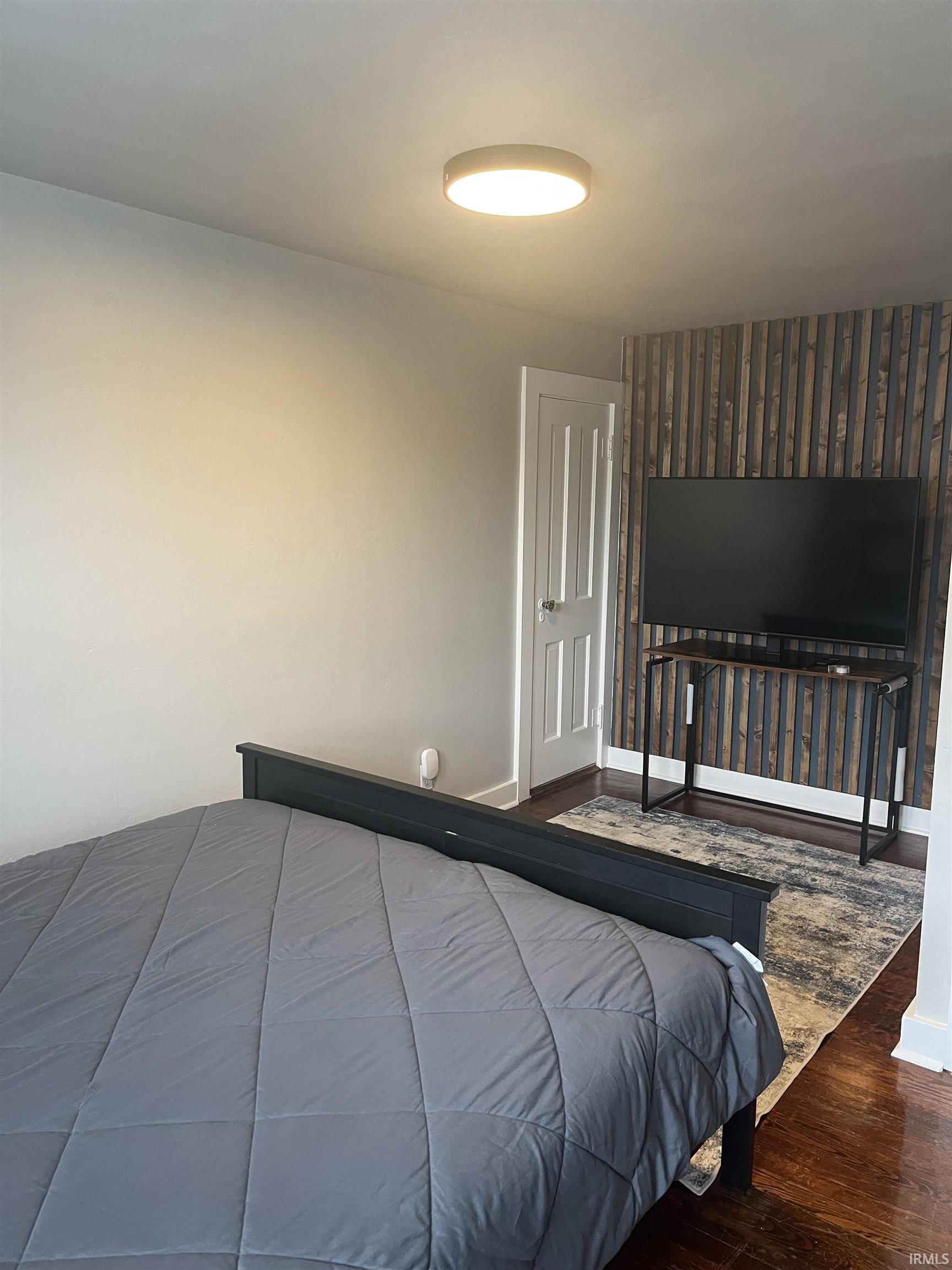 Bedroom featuring a decorative accent wall with vertical wood paneling, dark wood-finish flooring, a flush-mount ceiling light fixture, and a solid white door with panel detailing