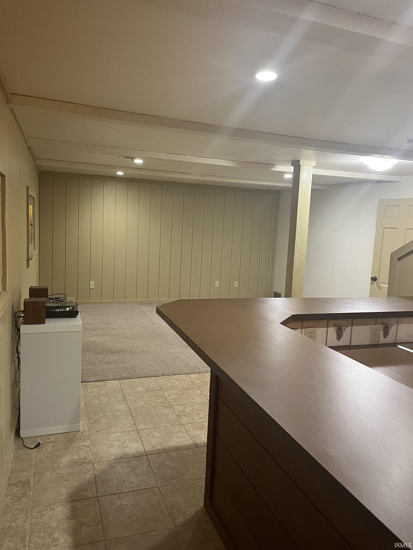 Recreational space featuring a built-in bar with a brown countertop, tiled flooring, recessed ceiling lighting, a light-colored wood-paneled accent wall, and a carpeted area