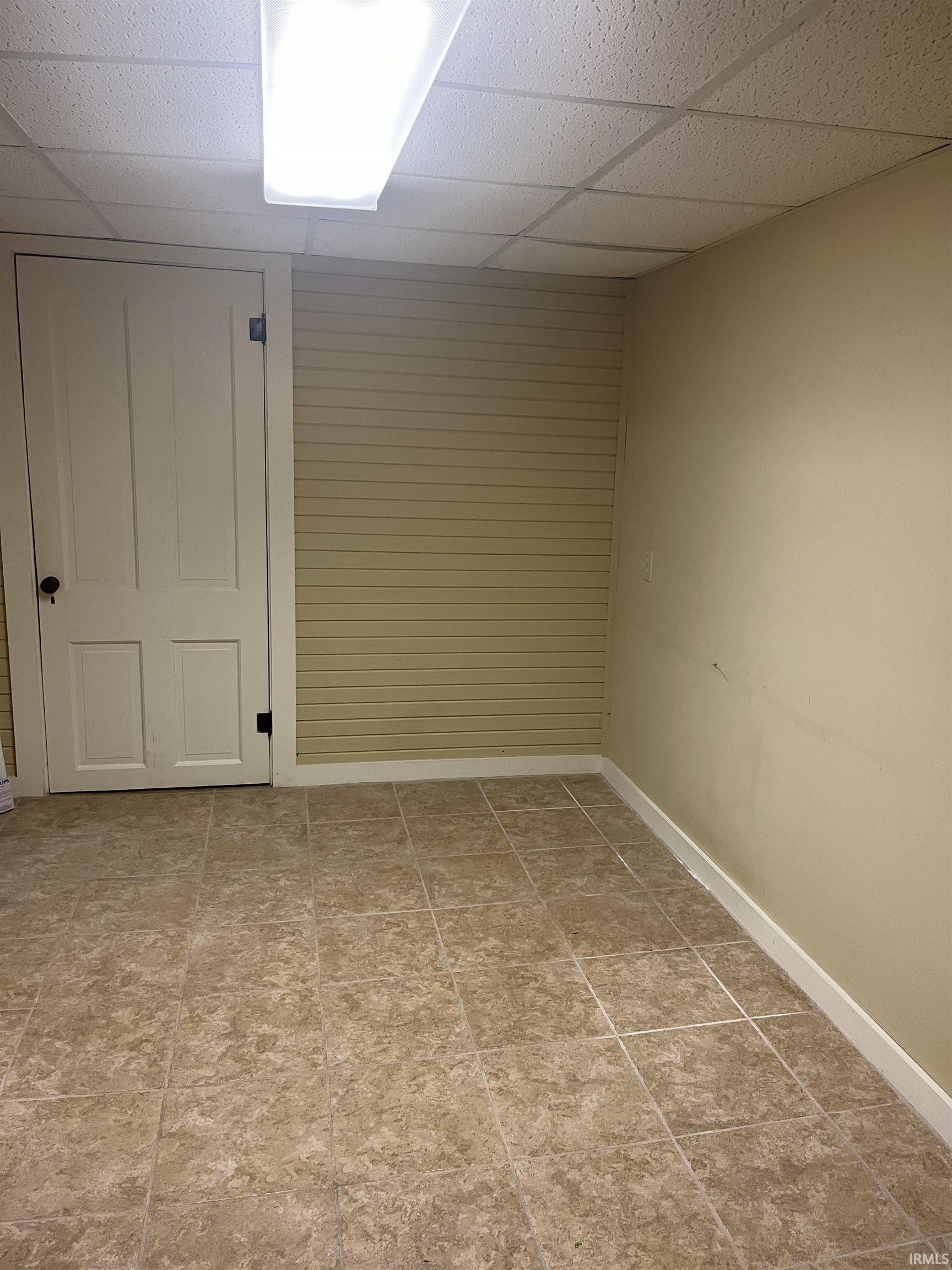 Interior room featuring square tan floor tiles, cream-colored walls, and a drop ceiling with a fluorescent light fixture
