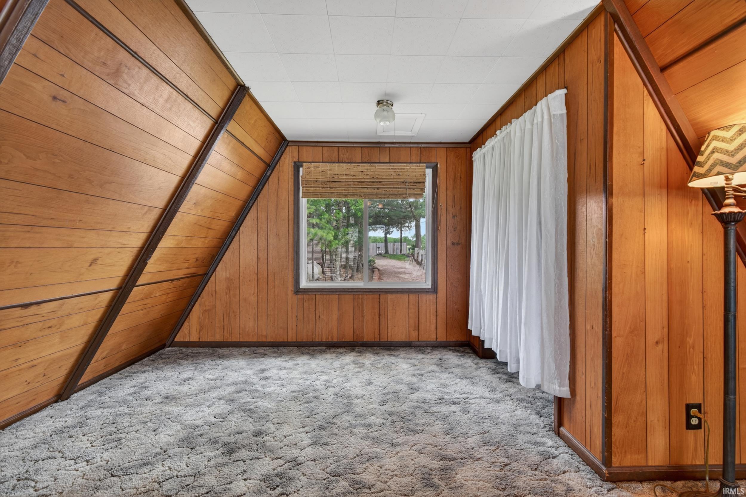 Interior space featuring wood paneling throughout, a large window with a natural woven shade, and plush gray carpet flooring