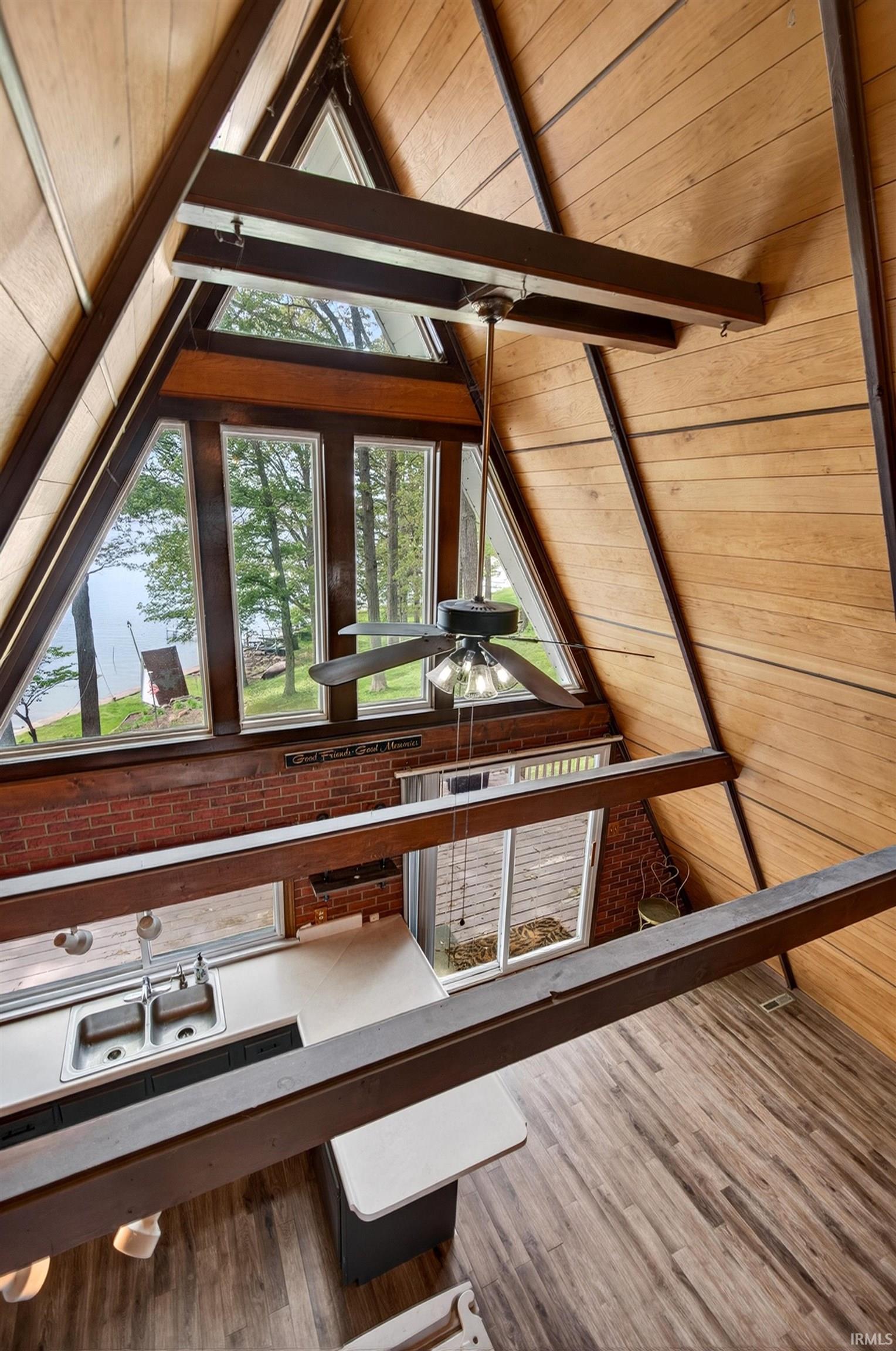 Vaulted wood-paneled ceilings with exposed beams, A-frame architectural design, wood-finish flooring, a kitchen area with a double basin stainless steel sink, and a brick accent wall with horizontal windows
