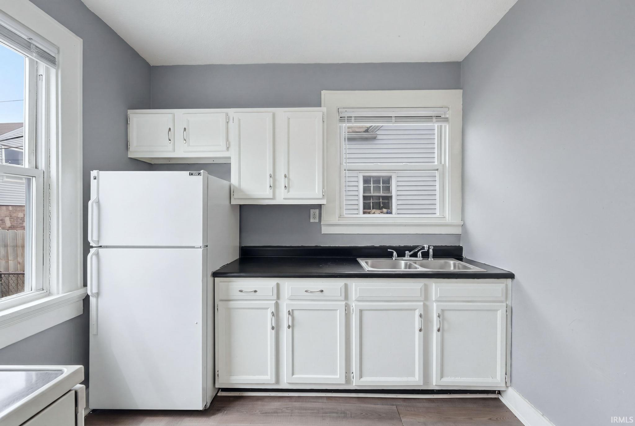 Kitchen space featuring white cabinetry, a double basin stainless steel sink, dark countertops, and a white refrigerator