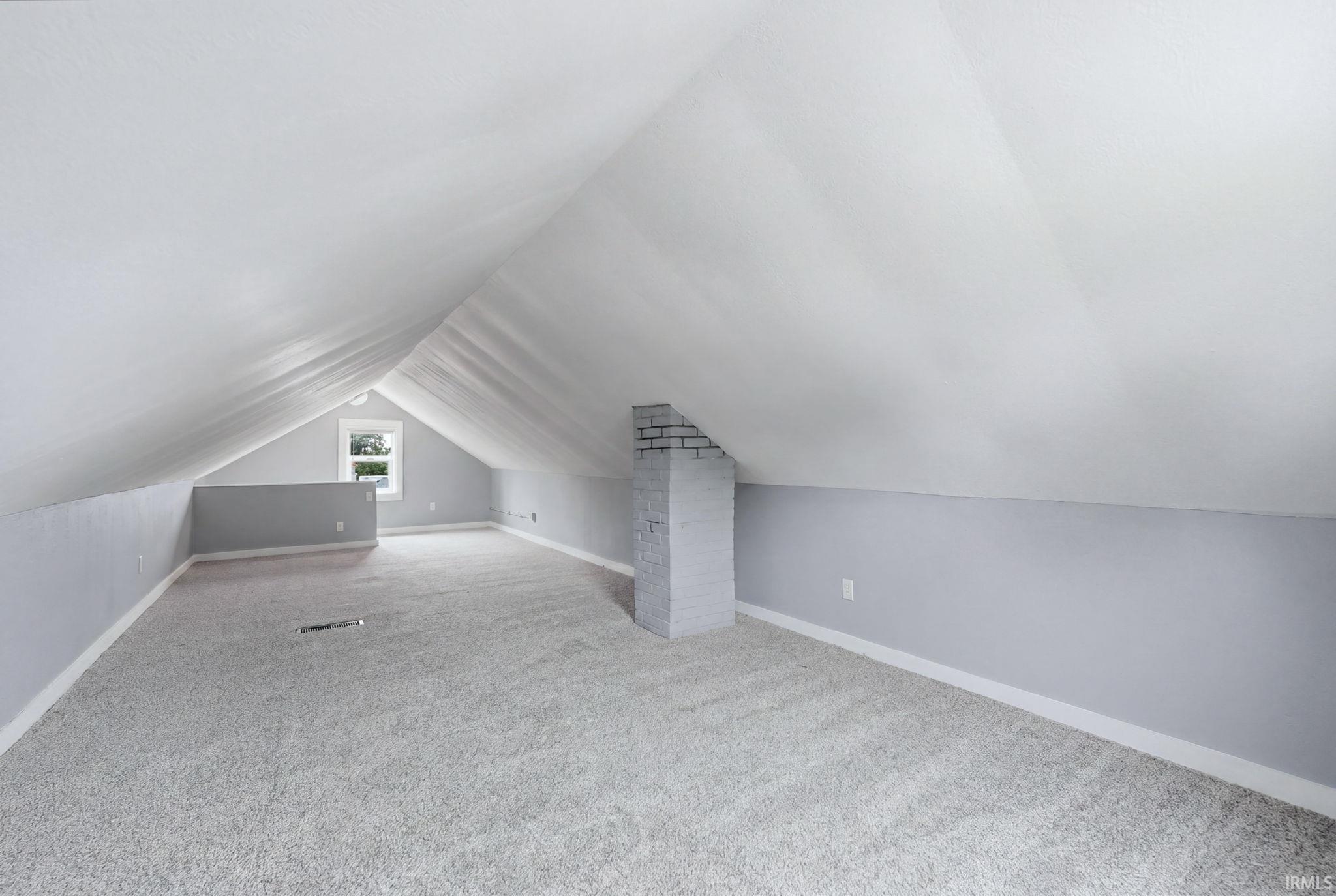 Expansive carpeted attic space featuring sloped ceilings, light gray walls, white trim, a double-hung window, and a brick-finish column
