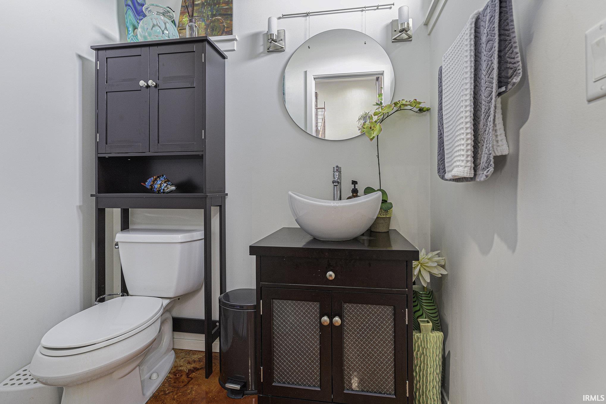 Contemporary bathroom featuring a vessel sink, dark wood-finish vanity with cabinet storage, circular wall mirror, and over-toilet storage cabinet