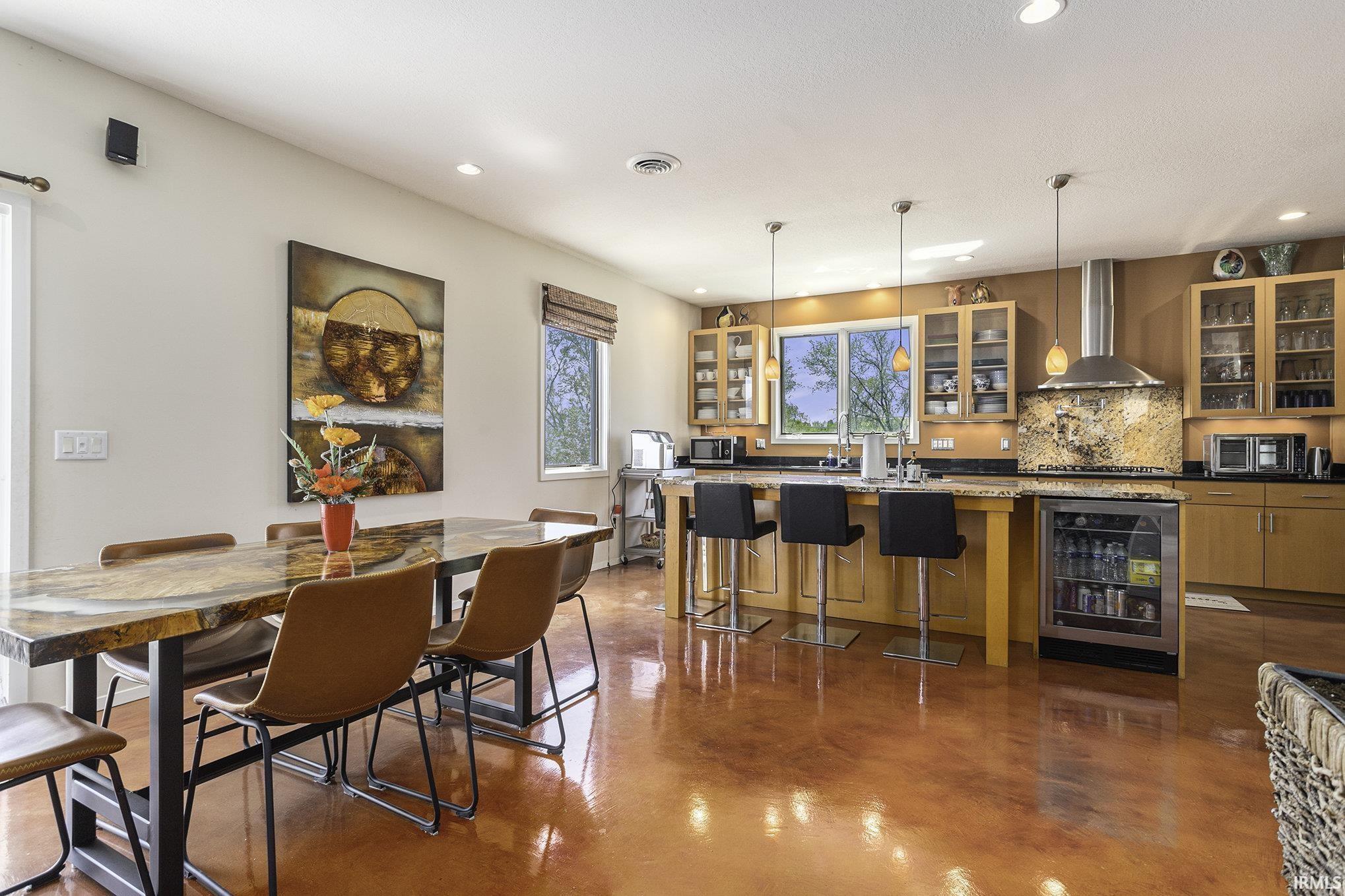 Open-concept living space featuring high-gloss terracotta-toned flooring, a spacious dining area with a stone-finish table, and a kitchen with amber-toned cabinetry, light-toned countertops, and a built-in beverage cooler