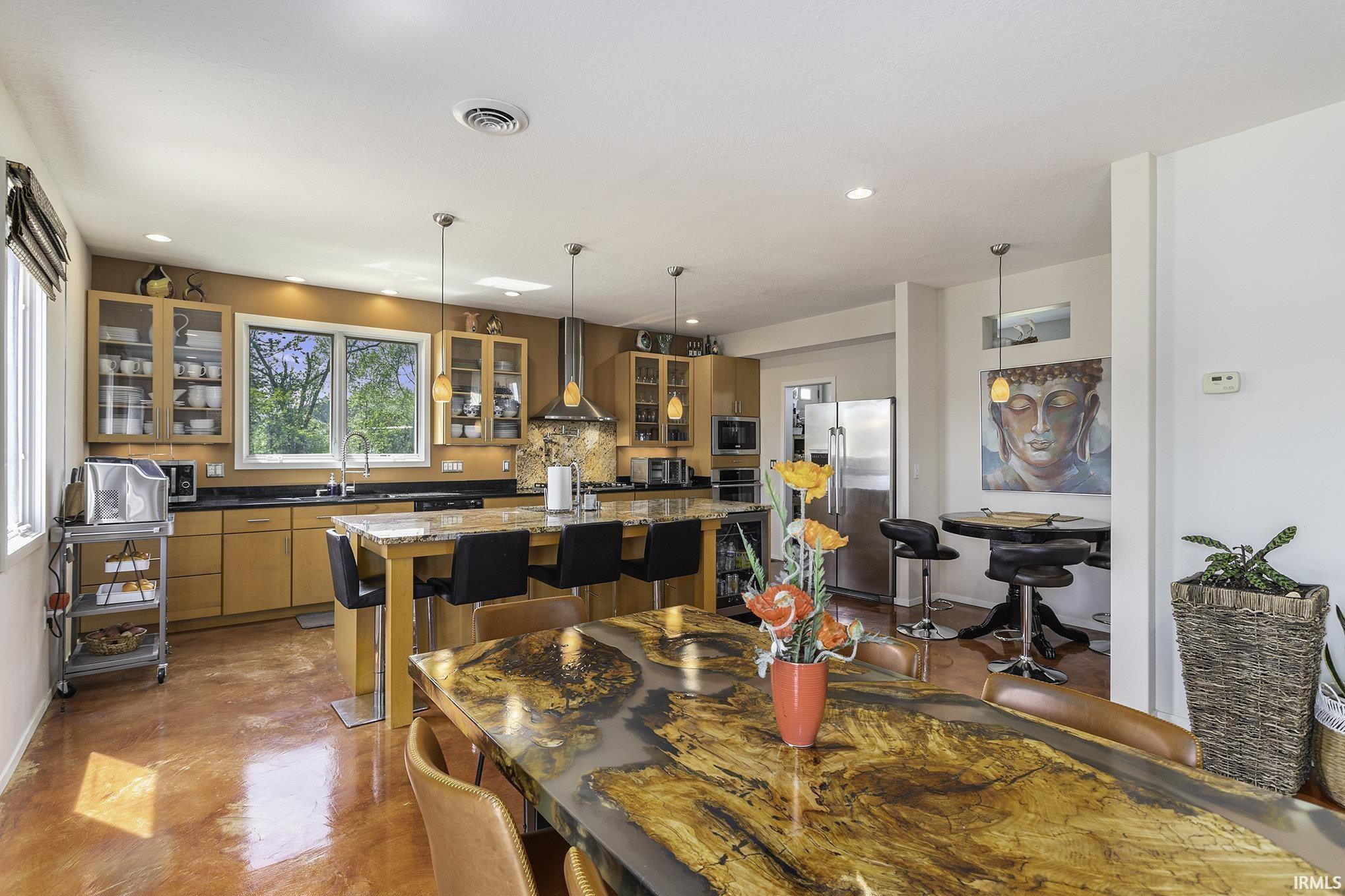 Open-concept kitchen and dining area featuring warm wood-finish cabinetry with glass inserts, dark countertops, and a full-height tile backsplash